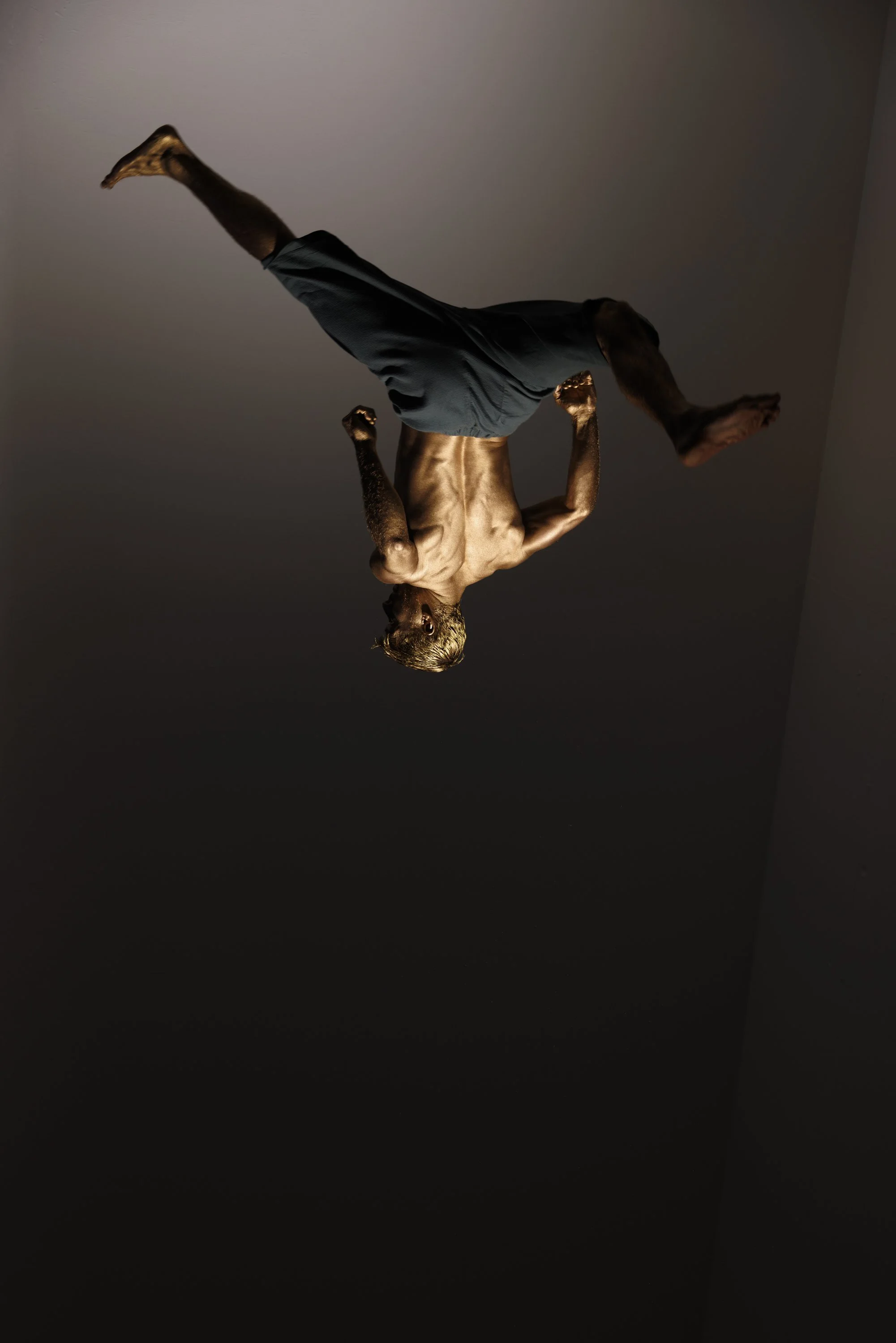 A person with a muscular build performing a handstand against a plain background. The person is shirtless, wearing dark shorts, and has a focused expression.