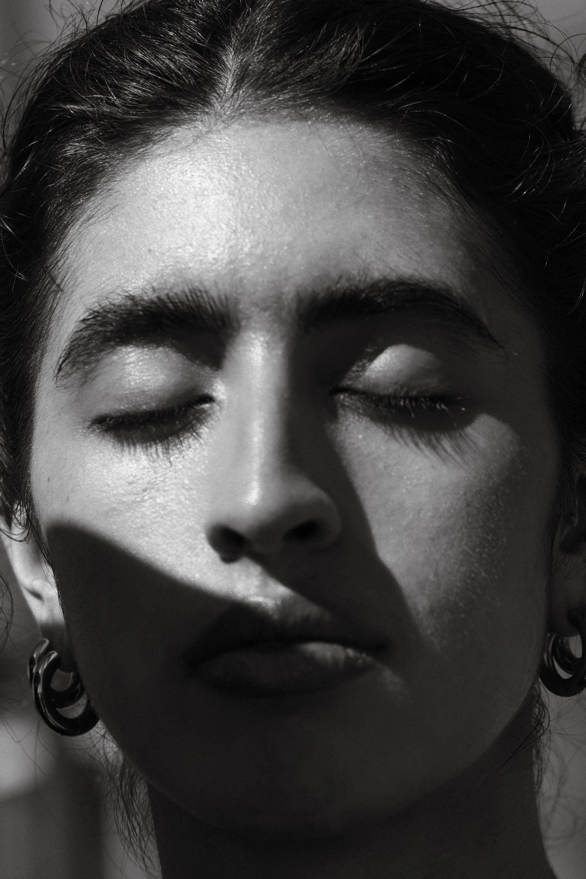 Close-up of a woman's face with eyes closed, high contrast black and white photo showing detailed skin texture and earrings.
