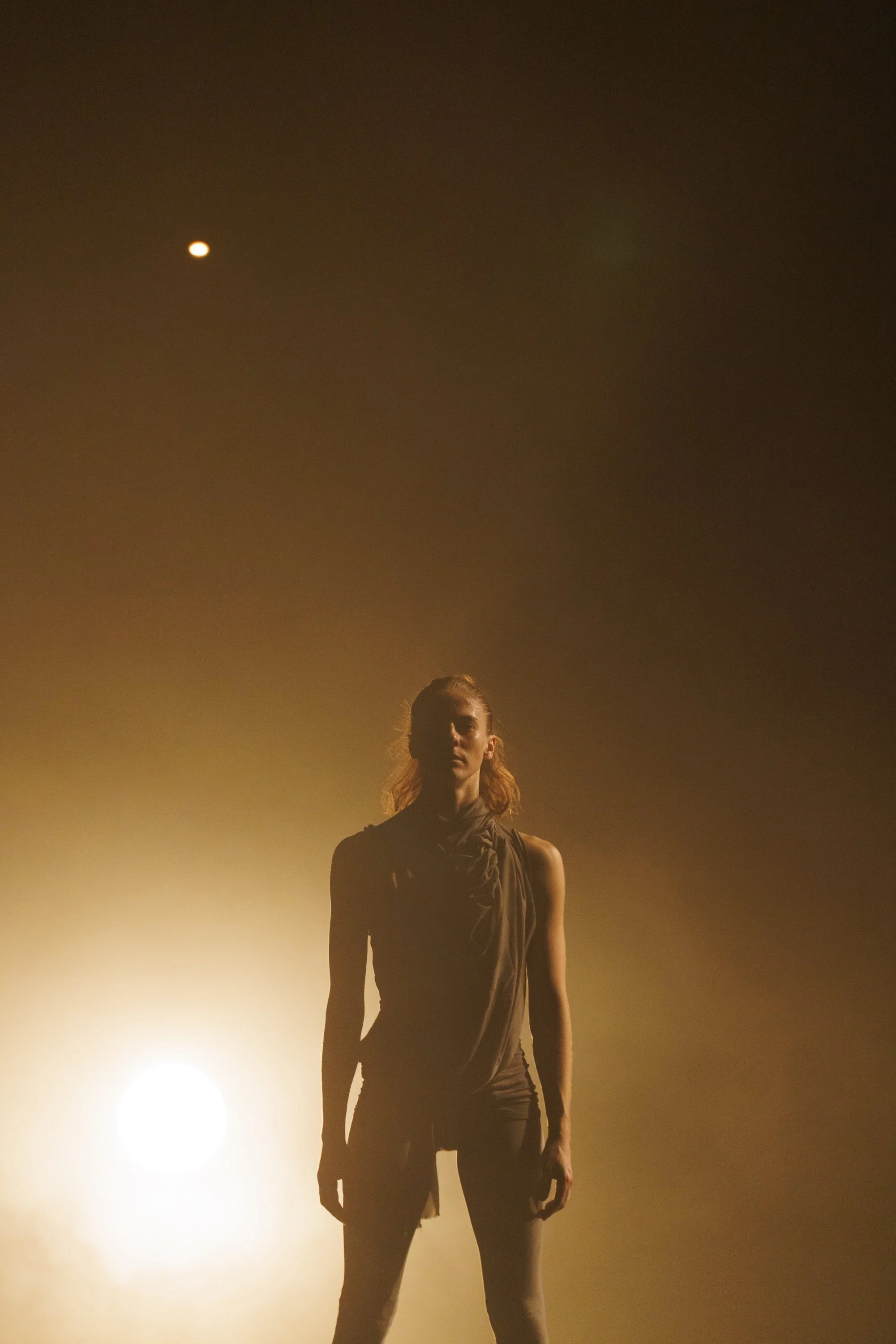 A woman standing in front of a bright light, with an orange glow and a dark sky with a visible moon above.