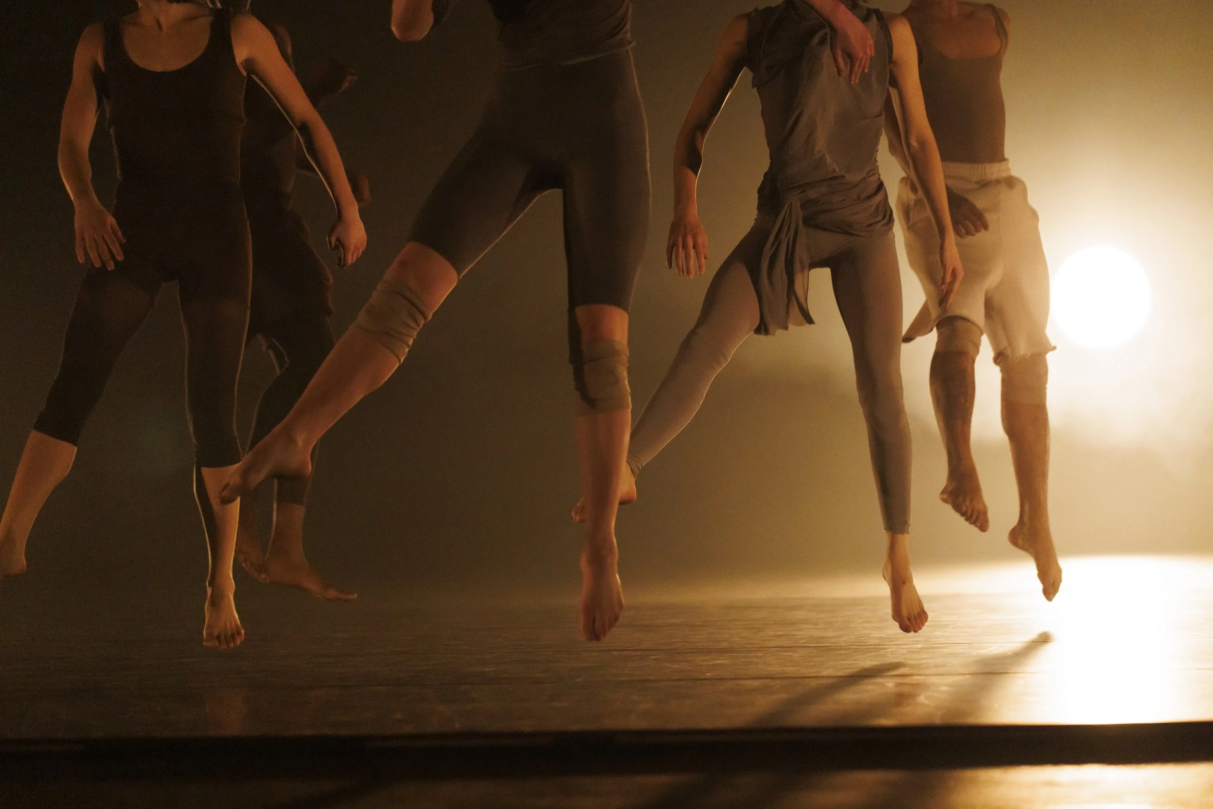 Dancers performing on stage, mid-air, with a bright light in the background creating a silhouette effect.