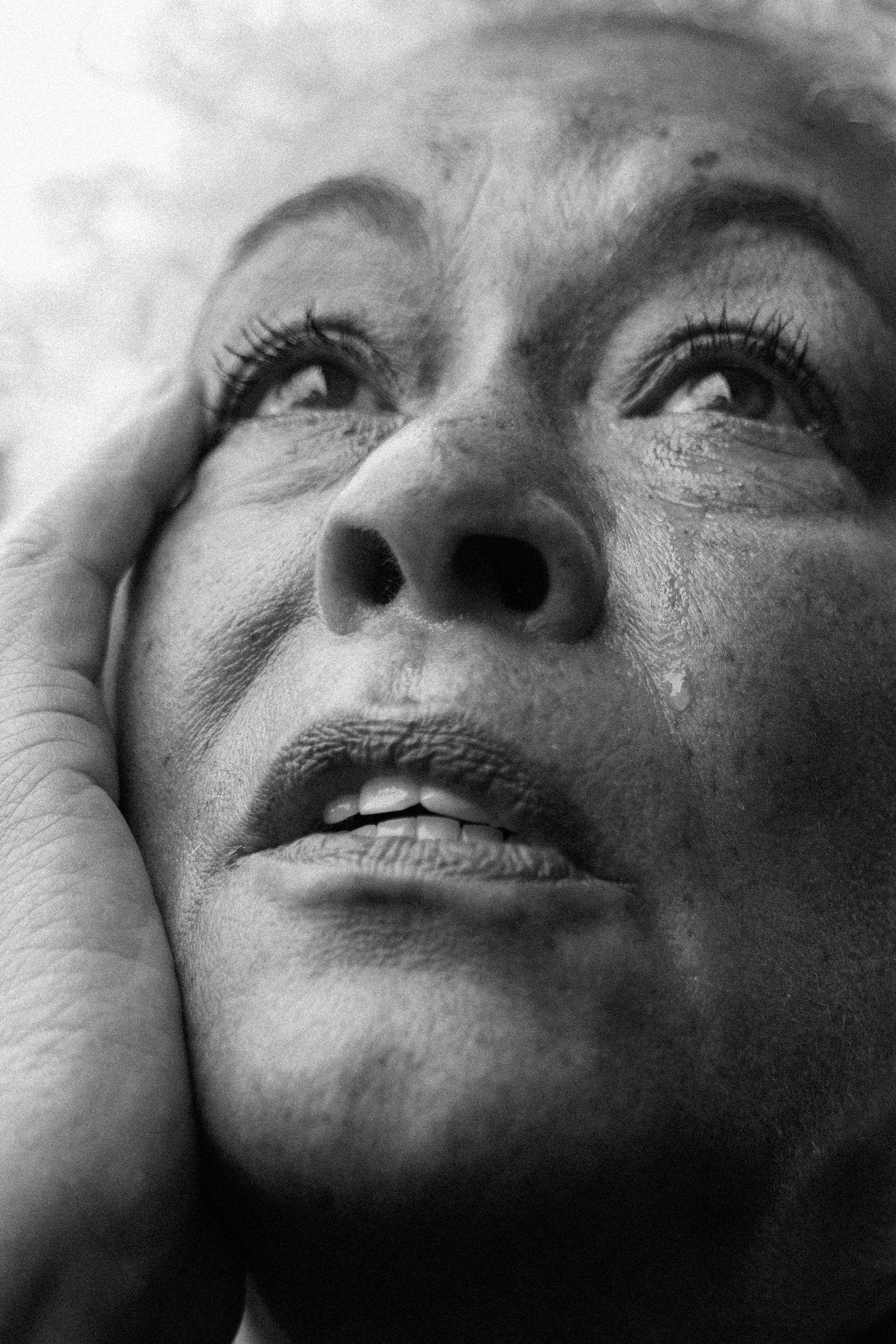 Close-up black and white photograph of a woman with a hand on her face, looking up with visible pores and textured skin.