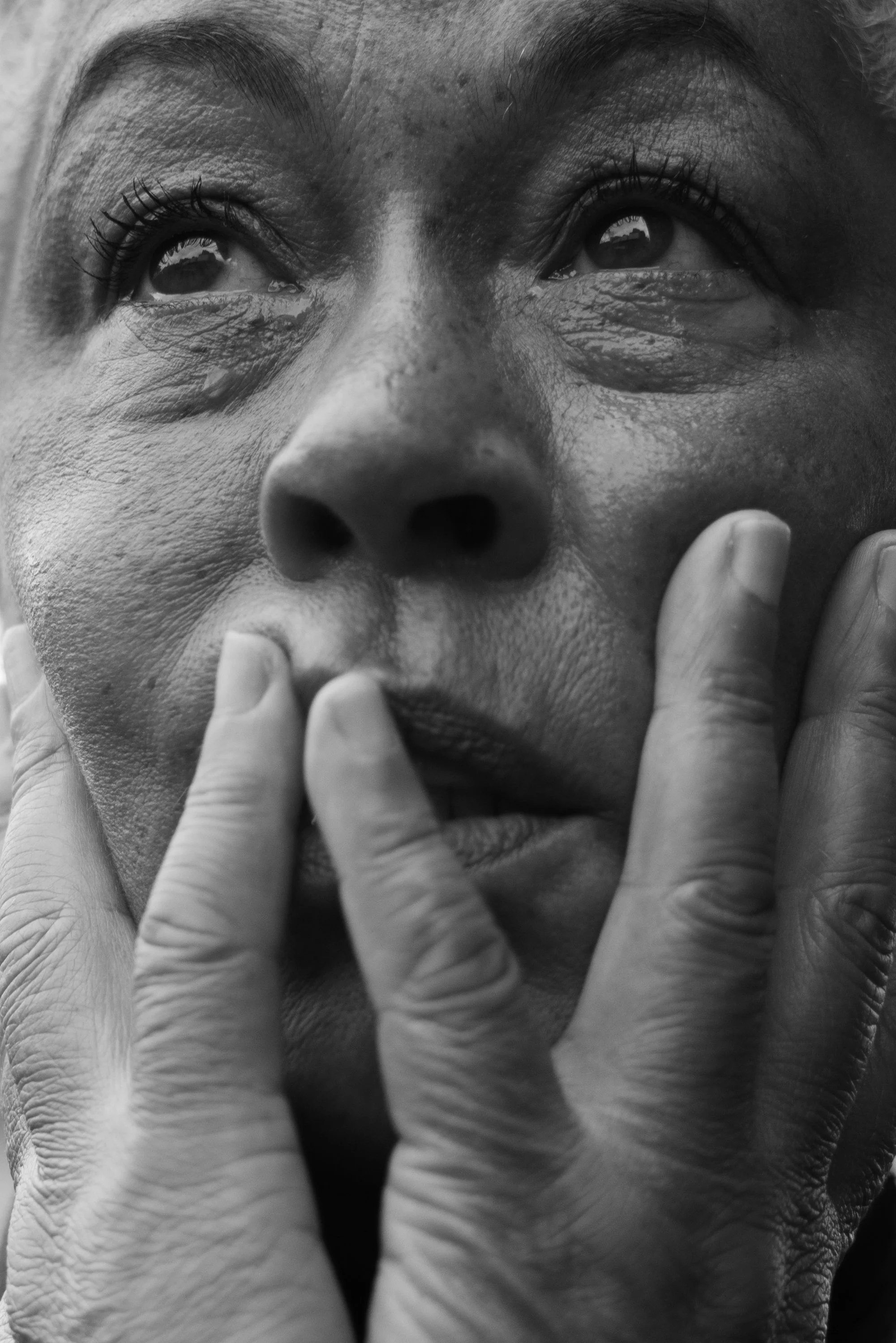 Close-up black and white portrait of a person's face with their fingers touching their lips, looking upward with expressive eyes.