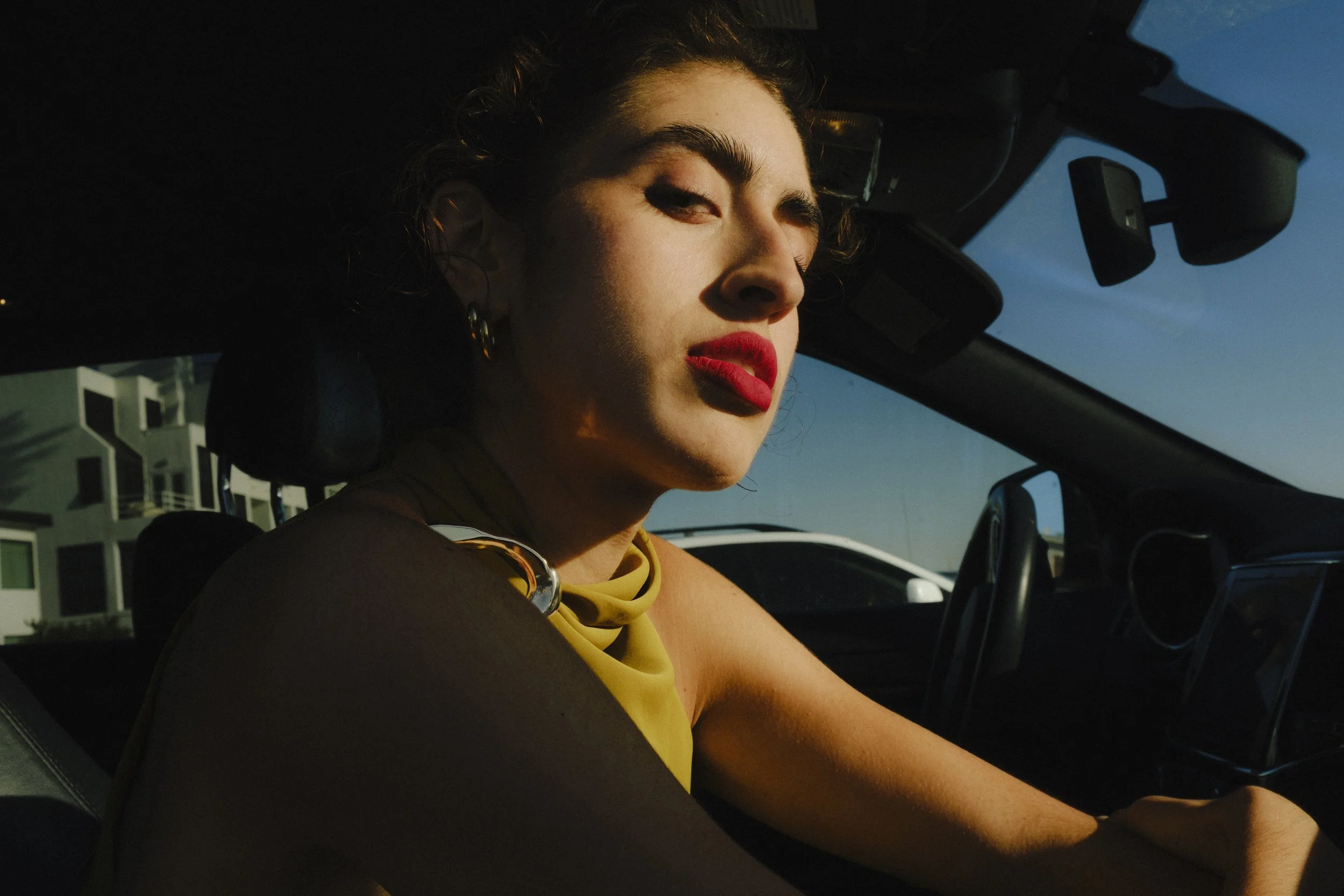 A woman with dark, curly hair, red lipstick, and earrings sitting in the driver's seat of a car, looking at the camera with a serious expression.