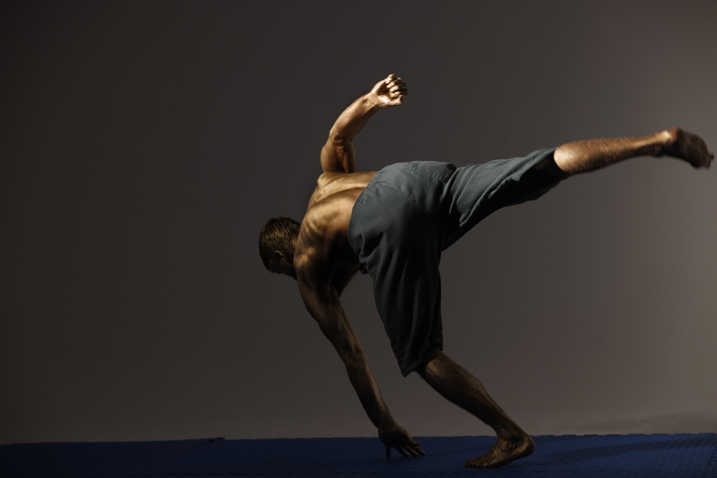 A shirtless man performing a complex balancing yoga pose on a blue mat against a neutral background.