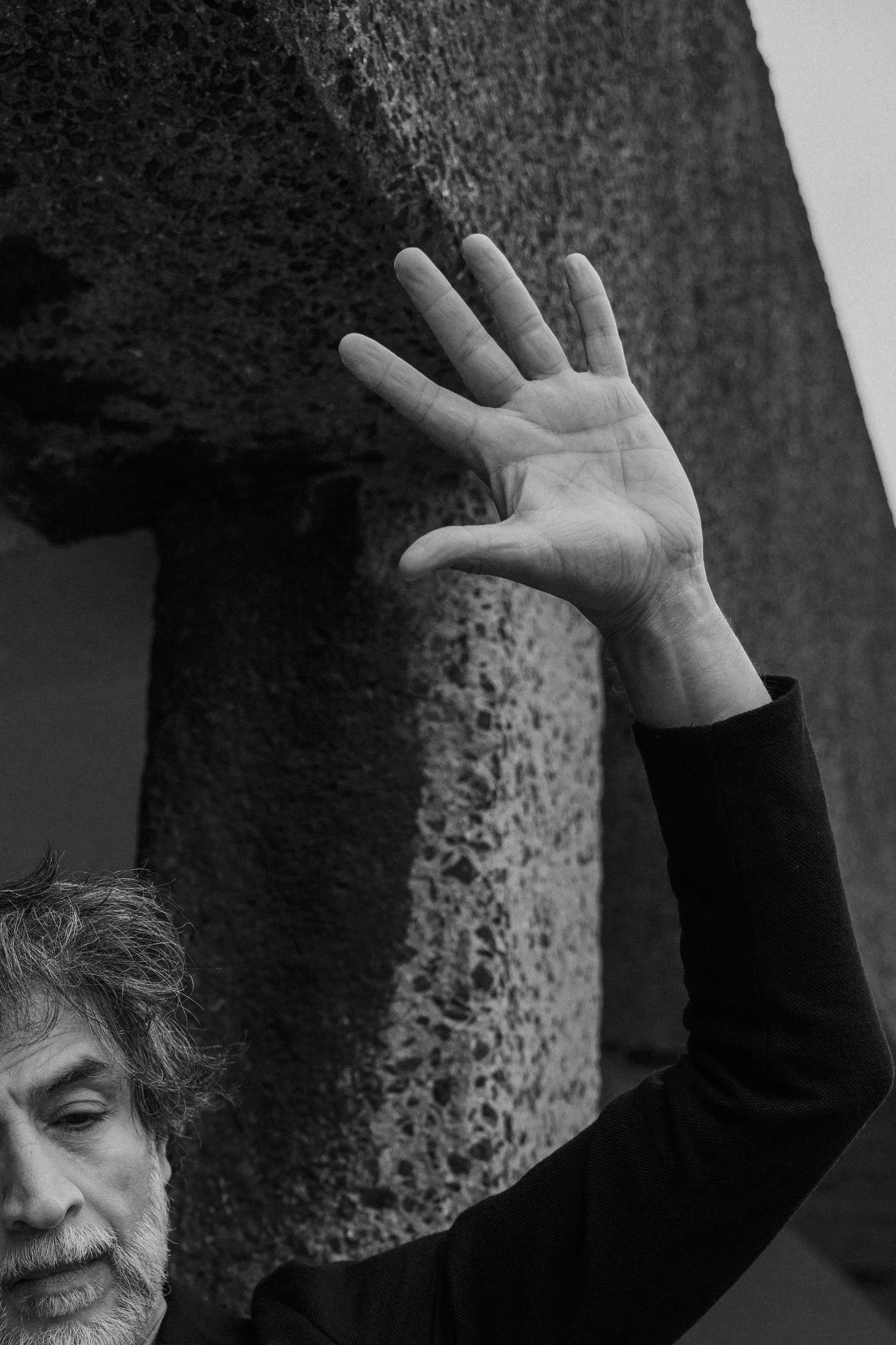 A man with gray hair and beard raising his right hand with fingers spread, against a textured vertical surface in a black and white photograph.