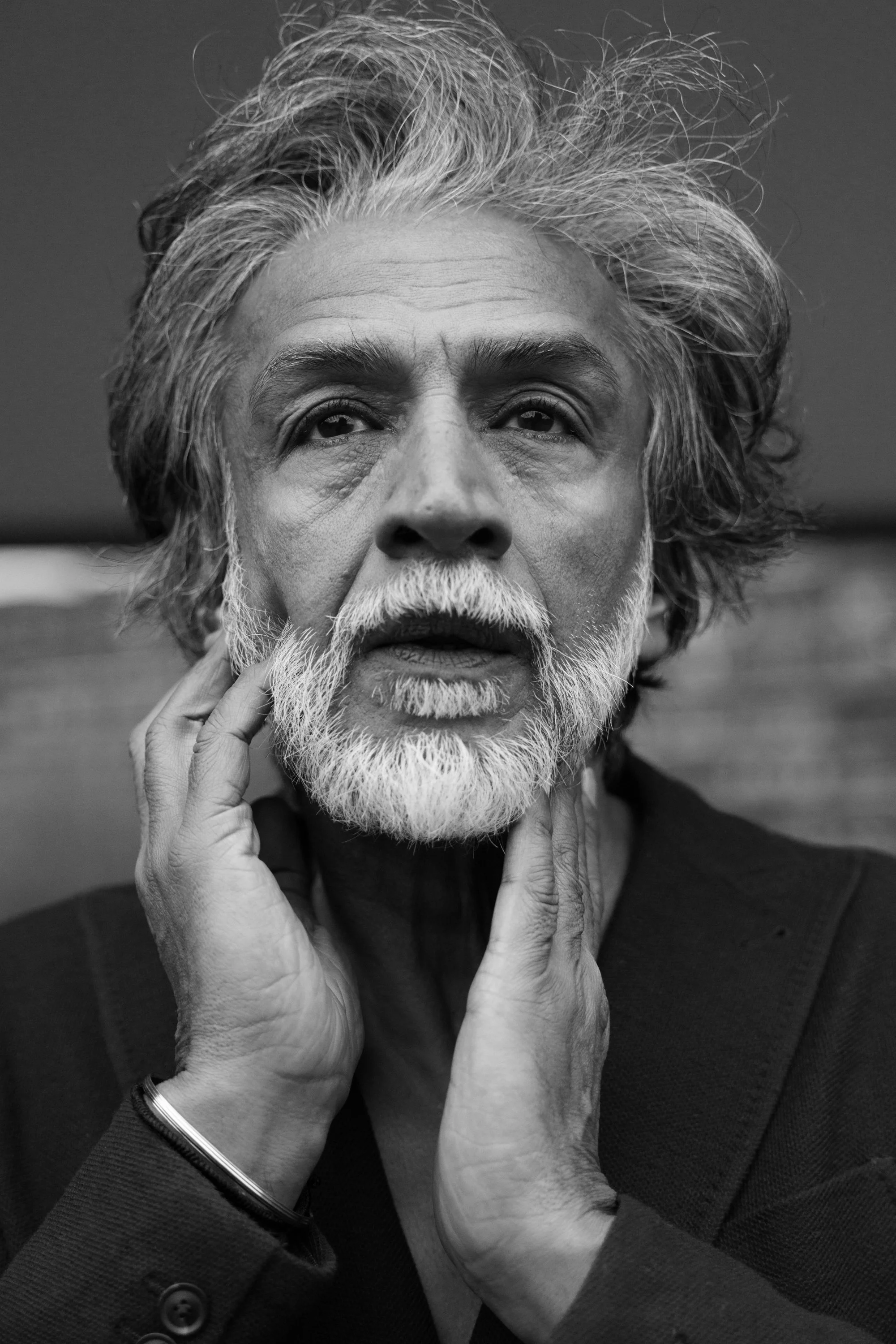 A black and white close-up portrait of an older man with a white beard and tousled hair, with his hands gently touching his face.
