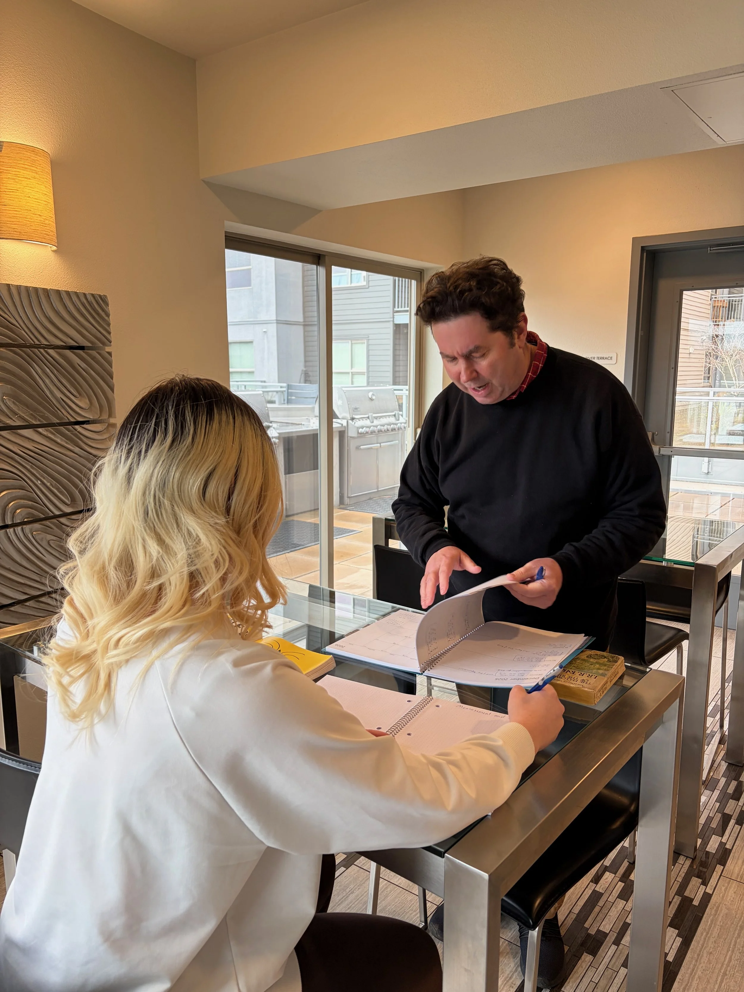 A woman with long blonde hair sitting at a desk, looking at a man standing and showing her a document, in a modern indoor setting with a glass door and outdoor patio.