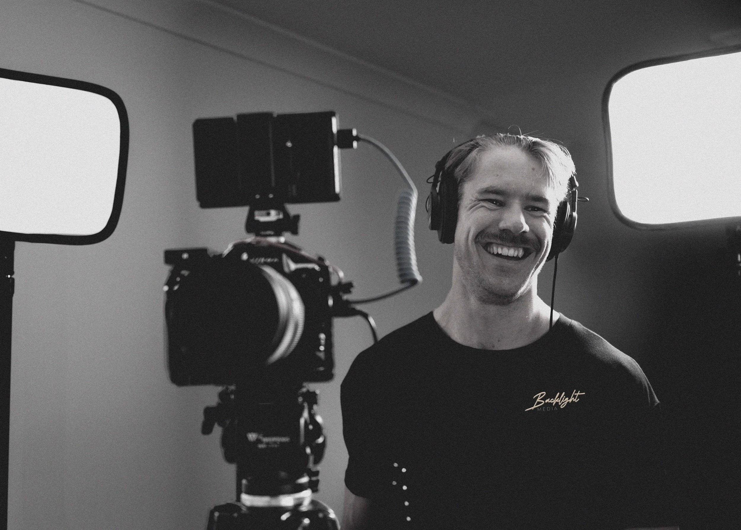 A young man with a mustache wearing headphones, smiling while being filmed or photographed in a studio with a camera on a tripod.