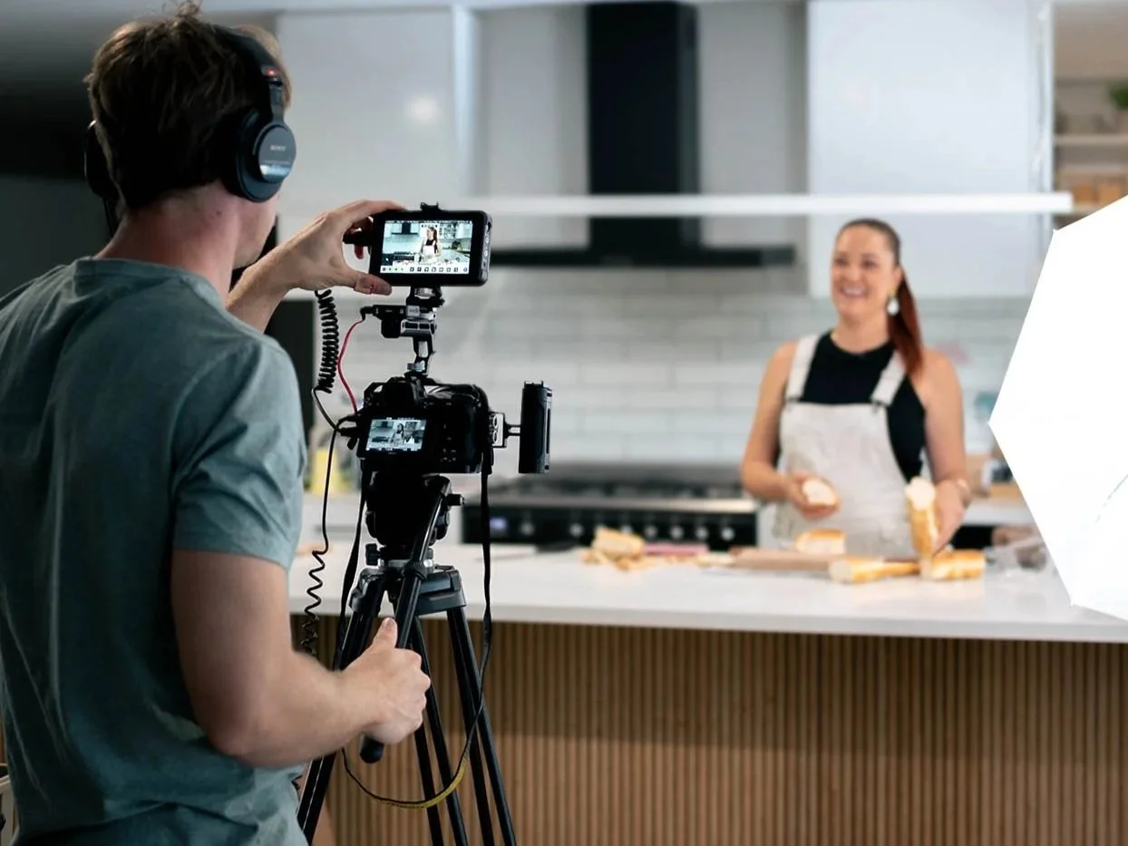 A camera operator wearing headphones is capturing a woman on set in a kitchen