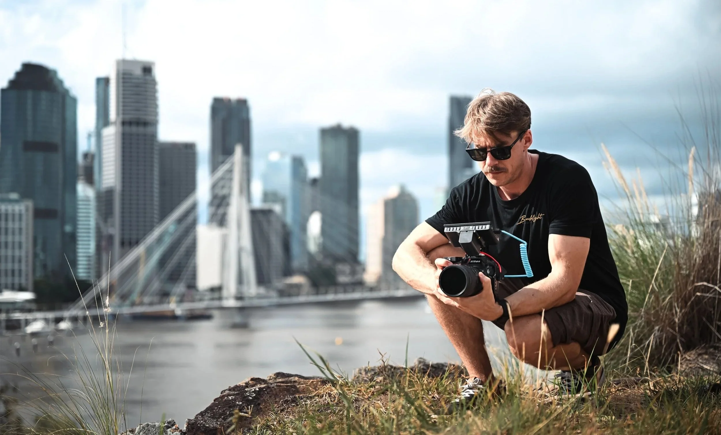 A man squatting outdoors on a grassy area with tall grass and rocks, holding a camera, with a city skyline and a bridge in the background.