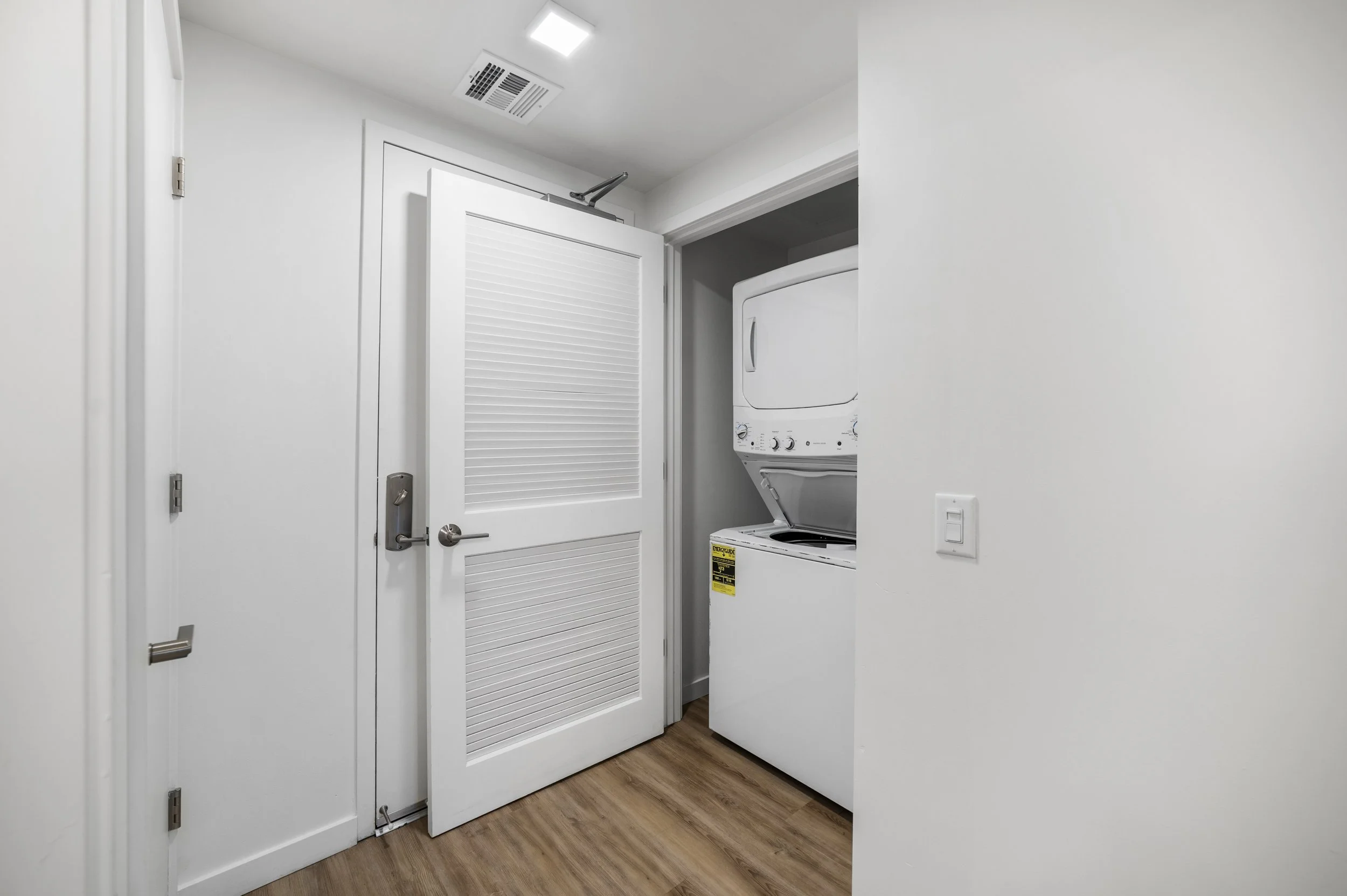 A laundry room with white walls, a wooden floor, and a white door slightly open revealing a stacked washer and dryer.