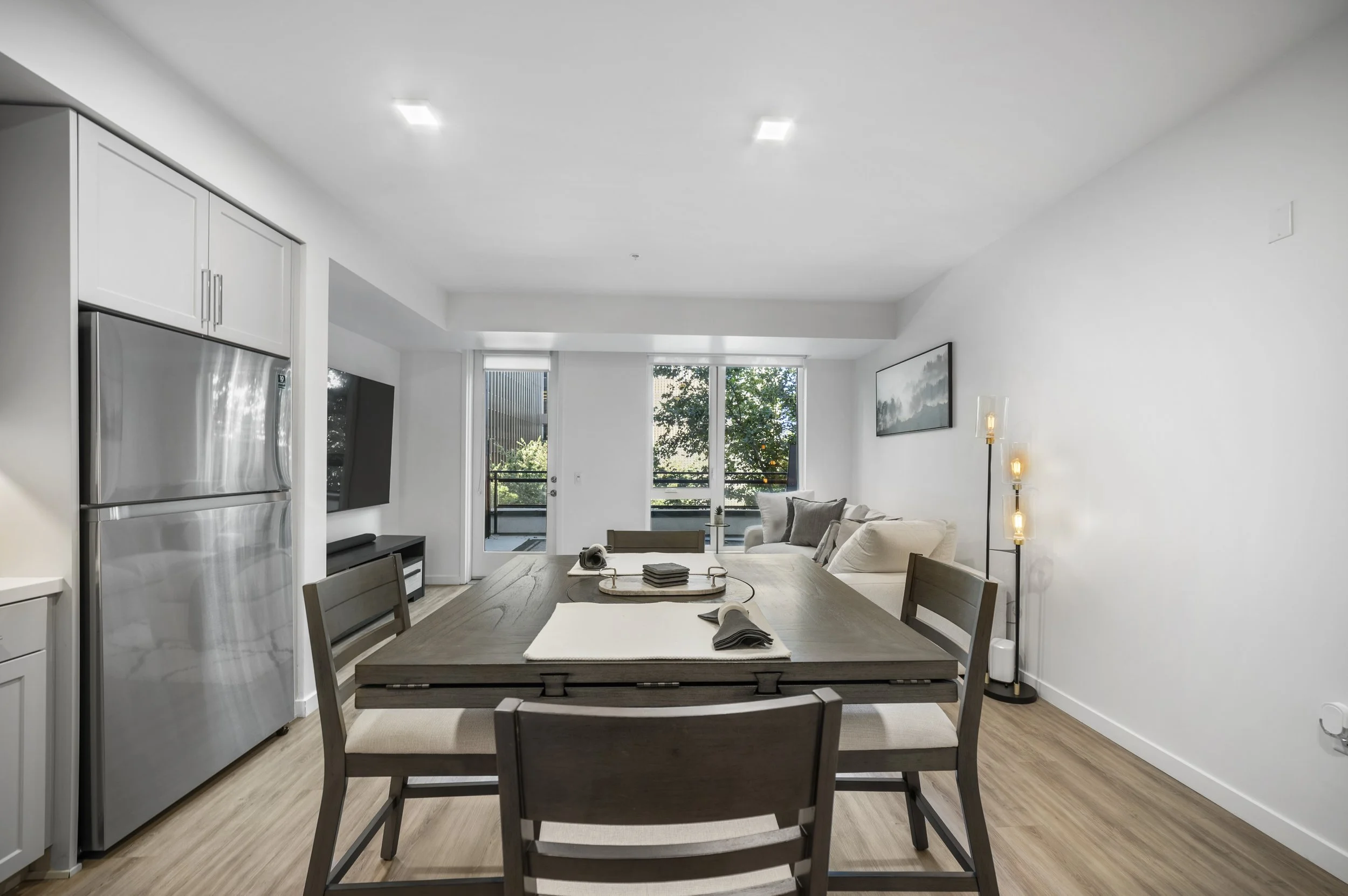 Modern open-concept living and dining area with a wooden dining table, beige chairs, a white sofa, wall art, large windows, and a balcony.