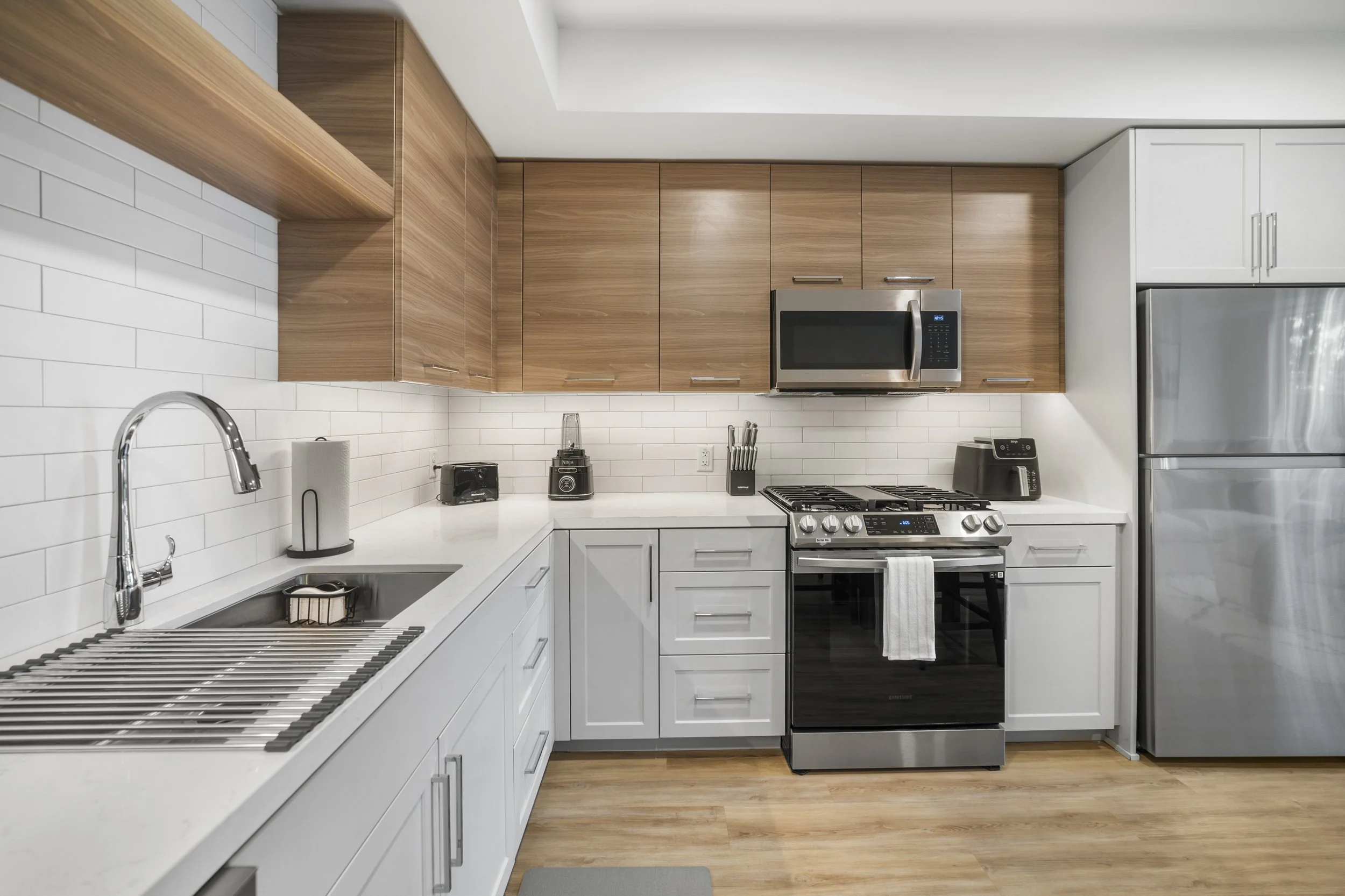 Modern kitchen with white cabinets, wooden upper cabinets, stainless steel appliances including a microwave, oven, toaster, blender, and refrigerator, and a white tiled backsplash.