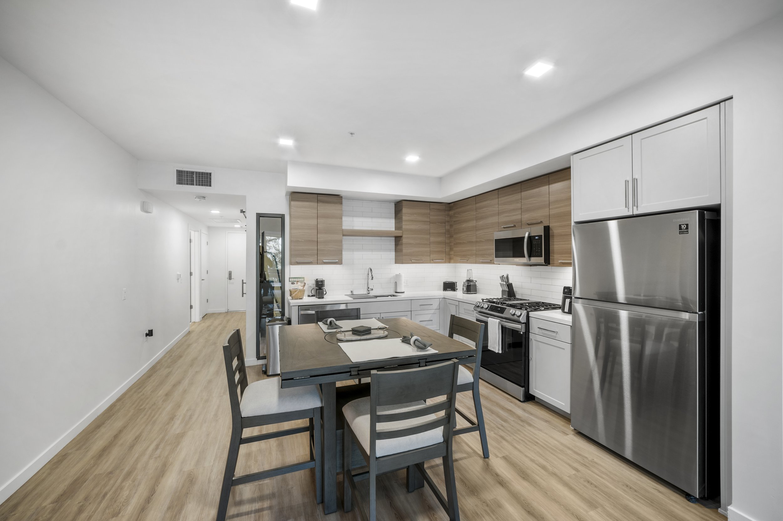 Modern kitchen with wooden cabinets, stainless steel appliances, a dining table with four chairs, white walls, and hardwood flooring.