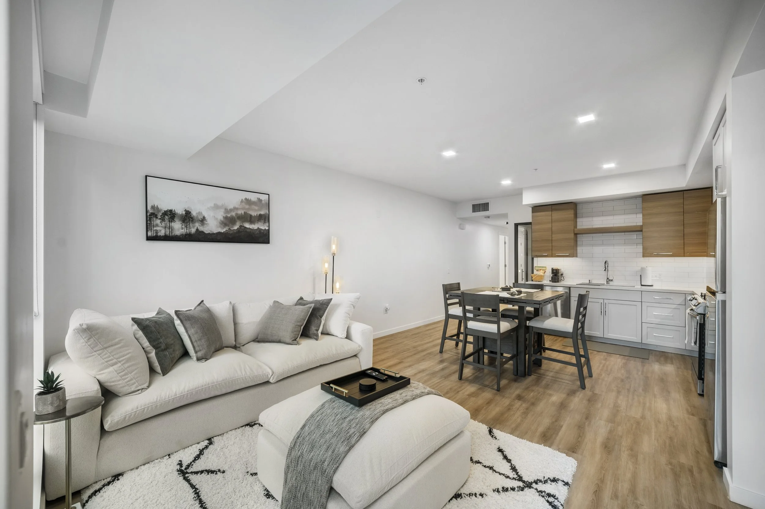 Modern open-concept living room and kitchen with white walls, light wood flooring, a white sofa with gray pillows, a black coffee table, a kitchen with wood cabinets, white backsplash, and a dining table with four chairs.