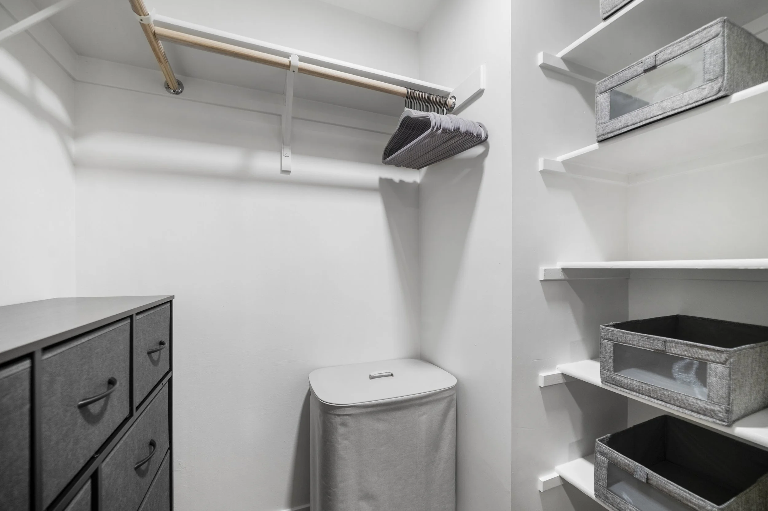 Empty walk-in closet with white walls and shelves, a wooden hanging rod with hangers, a small gray storage cabinet, and fabric bins on the shelves.