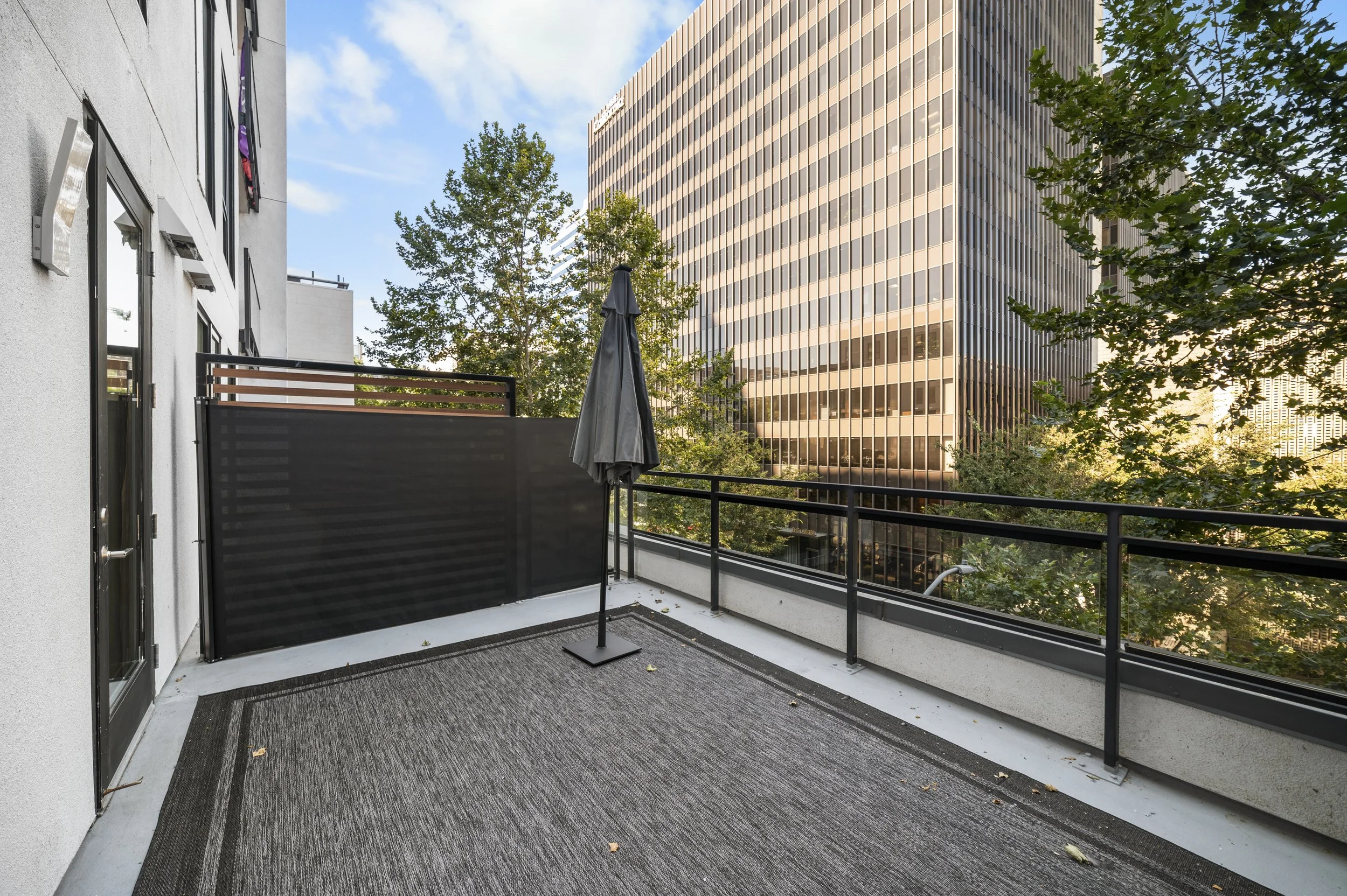 Empty balcony with black railing, grey rug, black umbrella, surrounded by tall city buildings and green trees.