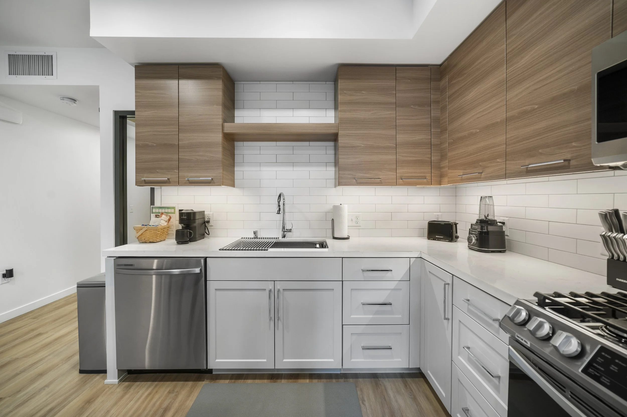 Modern kitchen with white lower cabinets, stainless steel appliances, wooden upper cabinets, white subway tile backsplash, and a gray rug on light wood flooring.