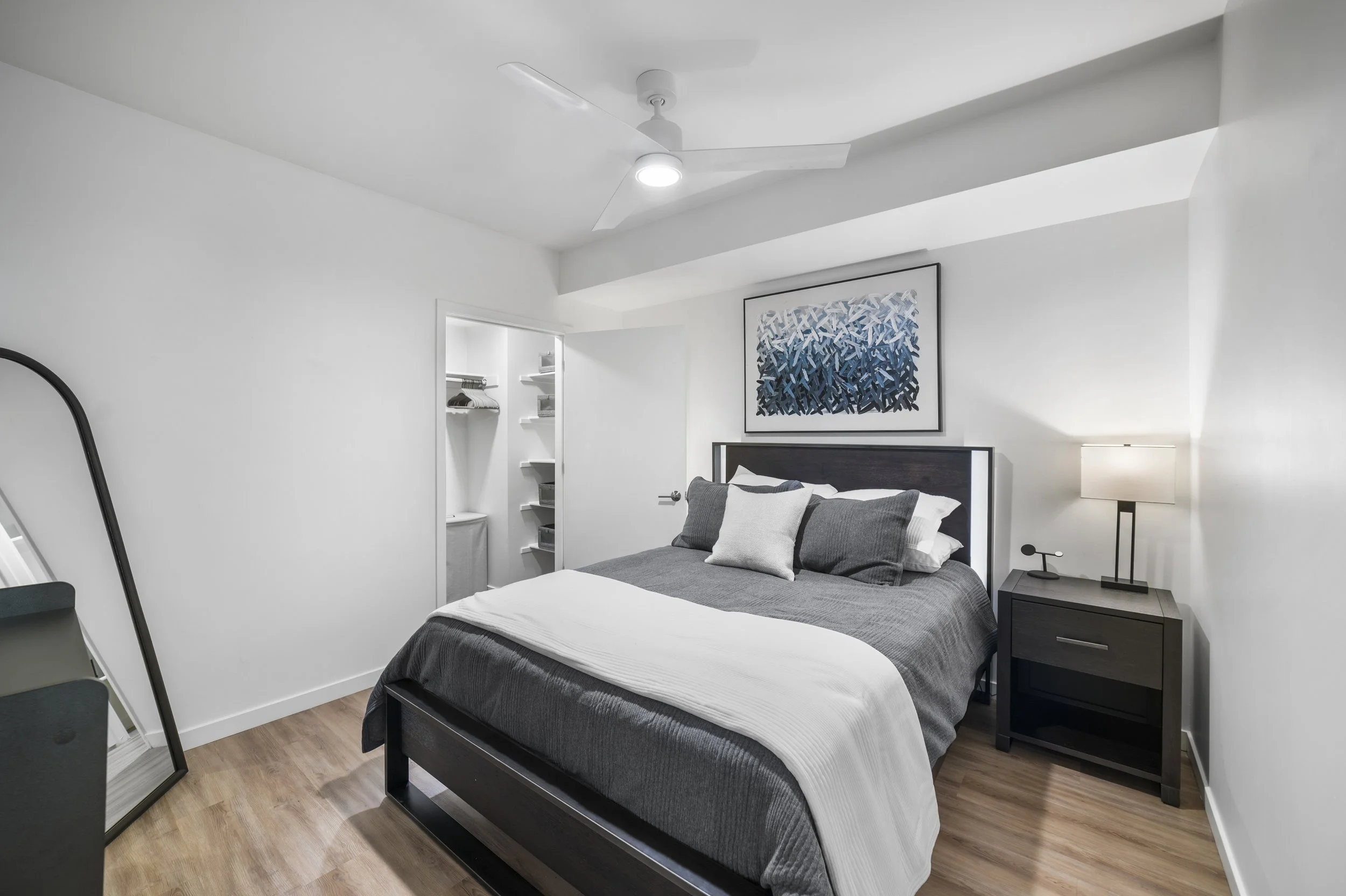 A modern bedroom with a bed, nightstand, lamp, and artwork, with a walk-in closet visible in the background.