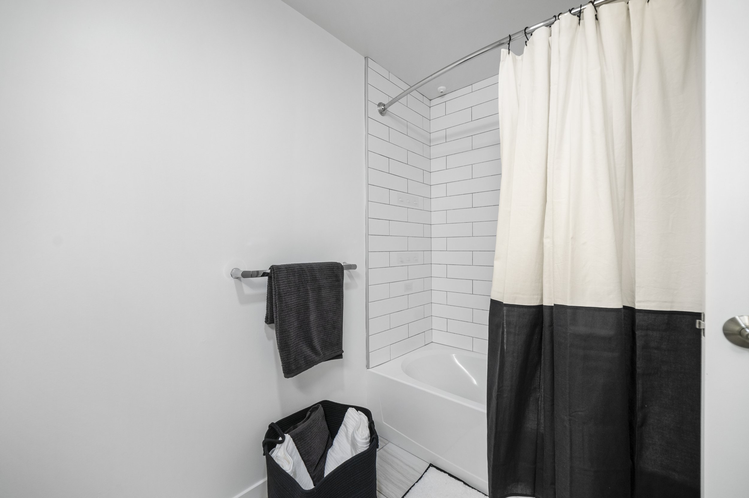 Empty bathroom with white walls, a corner bathtub, a shower curtain with black and white panels, a black towel on a silver towel bar, a black laundry basket with white towels, and light-colored wood flooring.