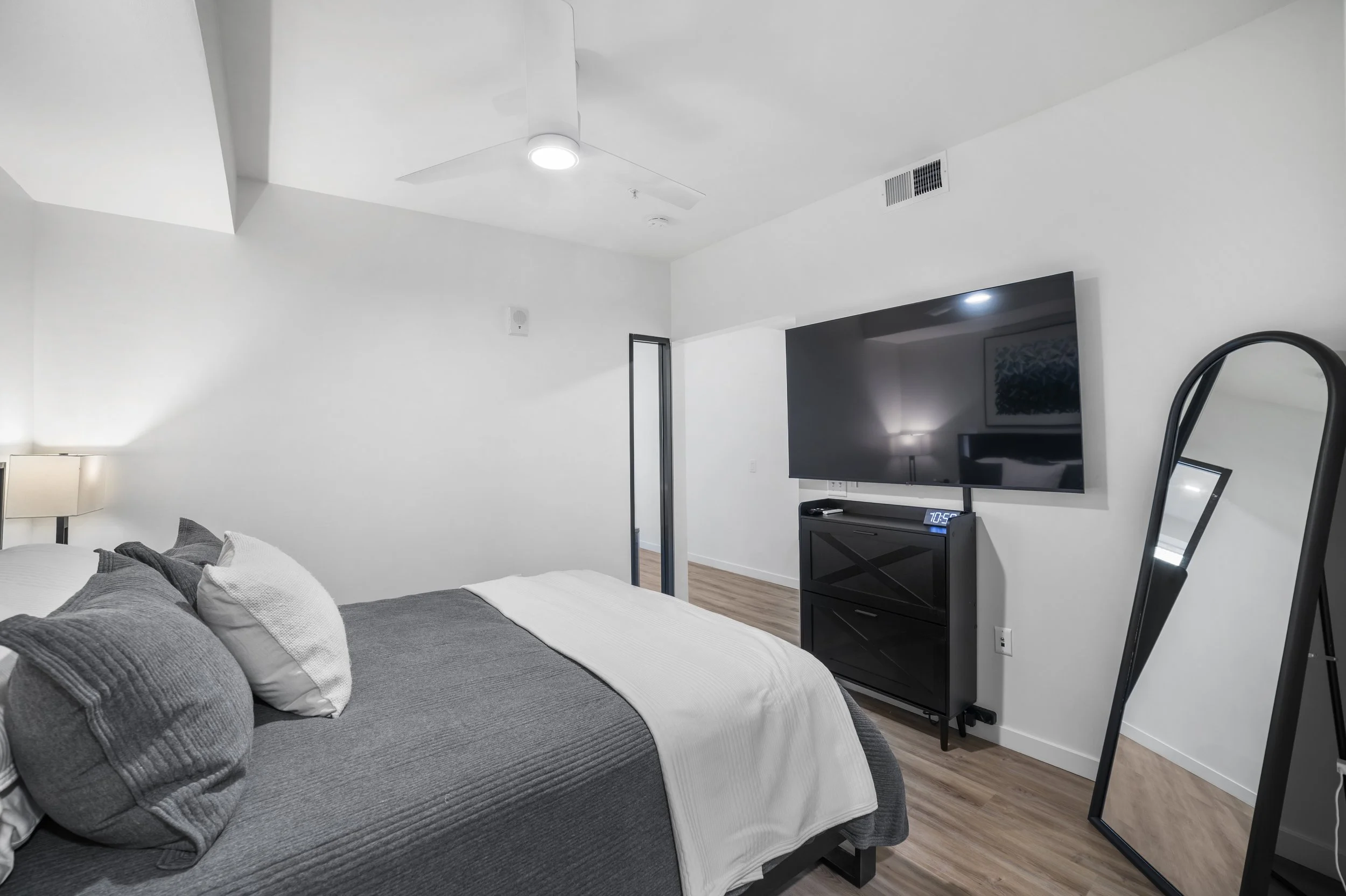Modern bedroom with white walls, a bed covered with gray and white bedding, a large flat-screen TV on a black stand, a full-length mirror, a ceiling fan, and wooden flooring.