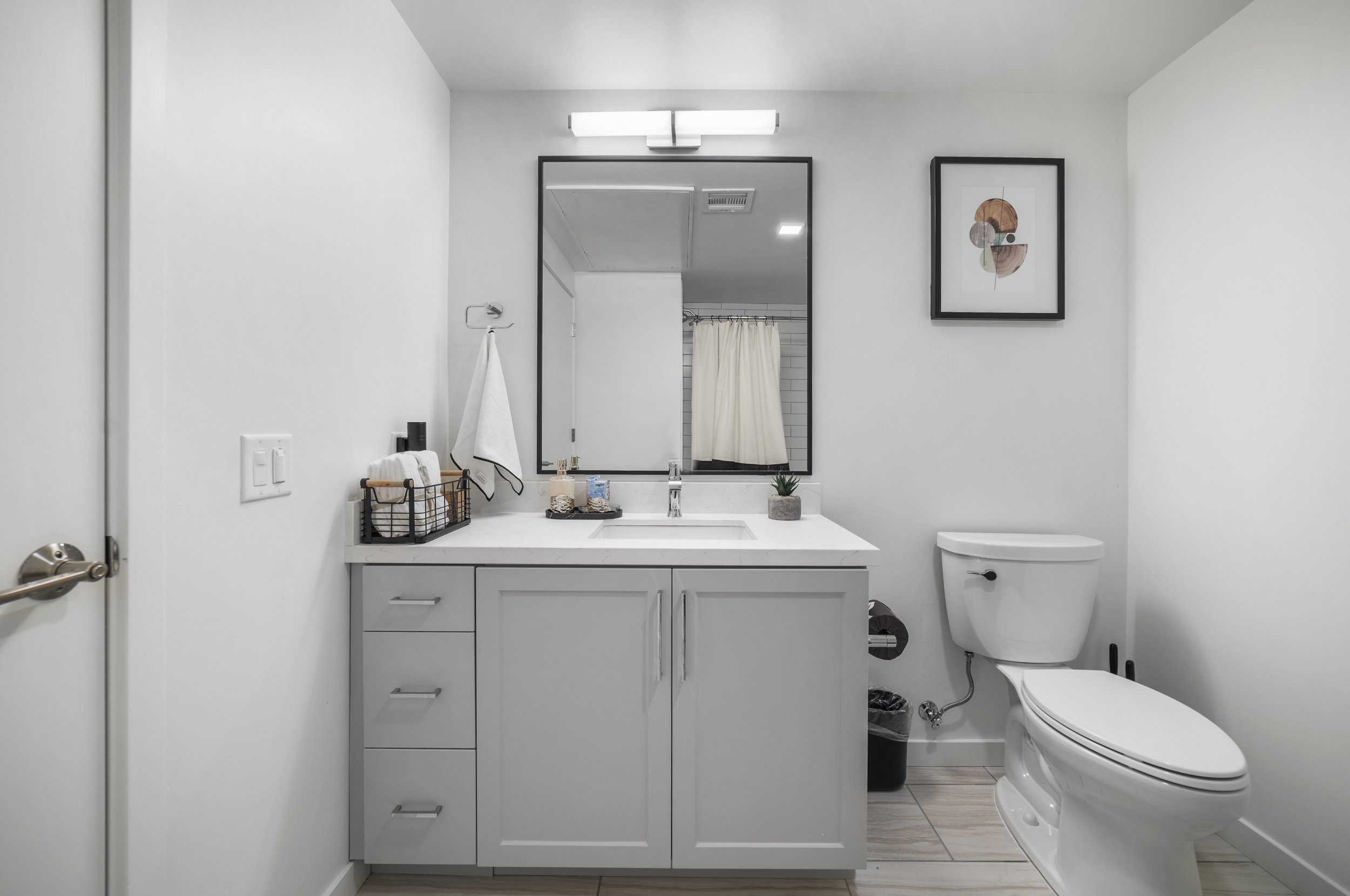 Modern bathroom with white vanity, black-framed mirror, artwork on wall, white toilet, and minimal decor.