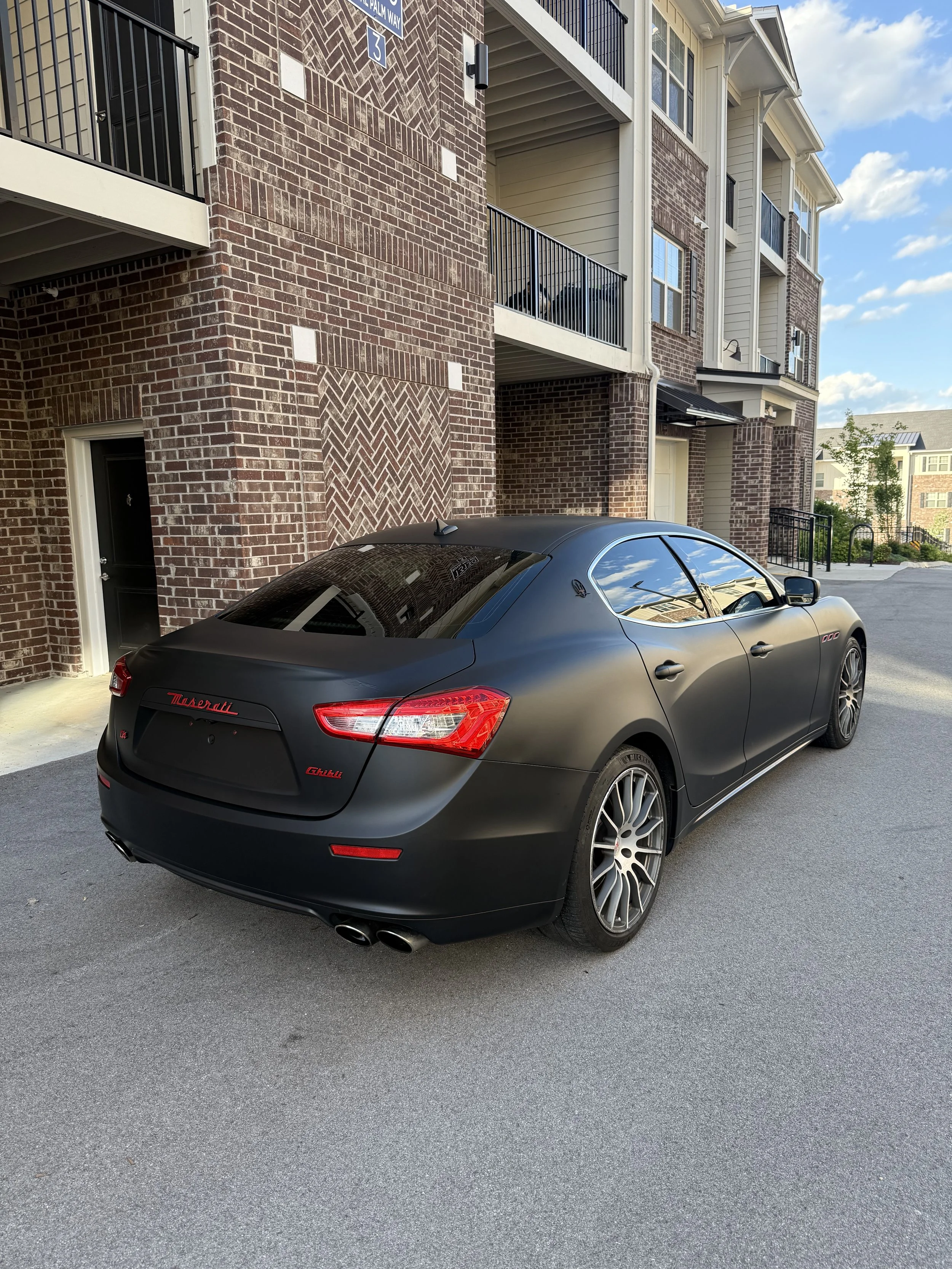 A black Maserati Ghibli parked outside a modern apartment building with brick and siding exterior.