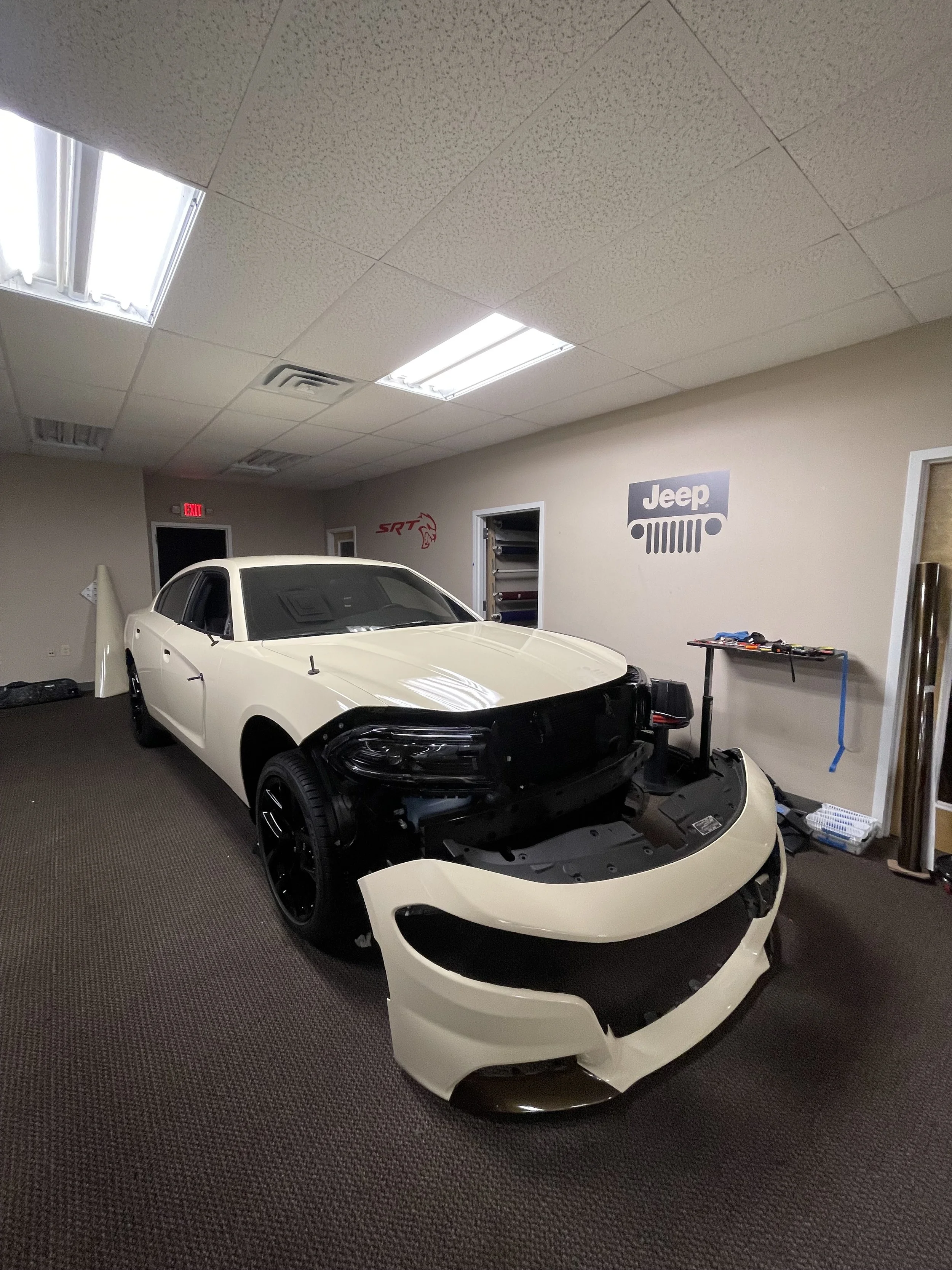 A white car in a garage or workshop, partially disassembled, with the front bumper and hood removed, surrounded by tools and automotive supplies, with Jeep and SRT logos on the wall.