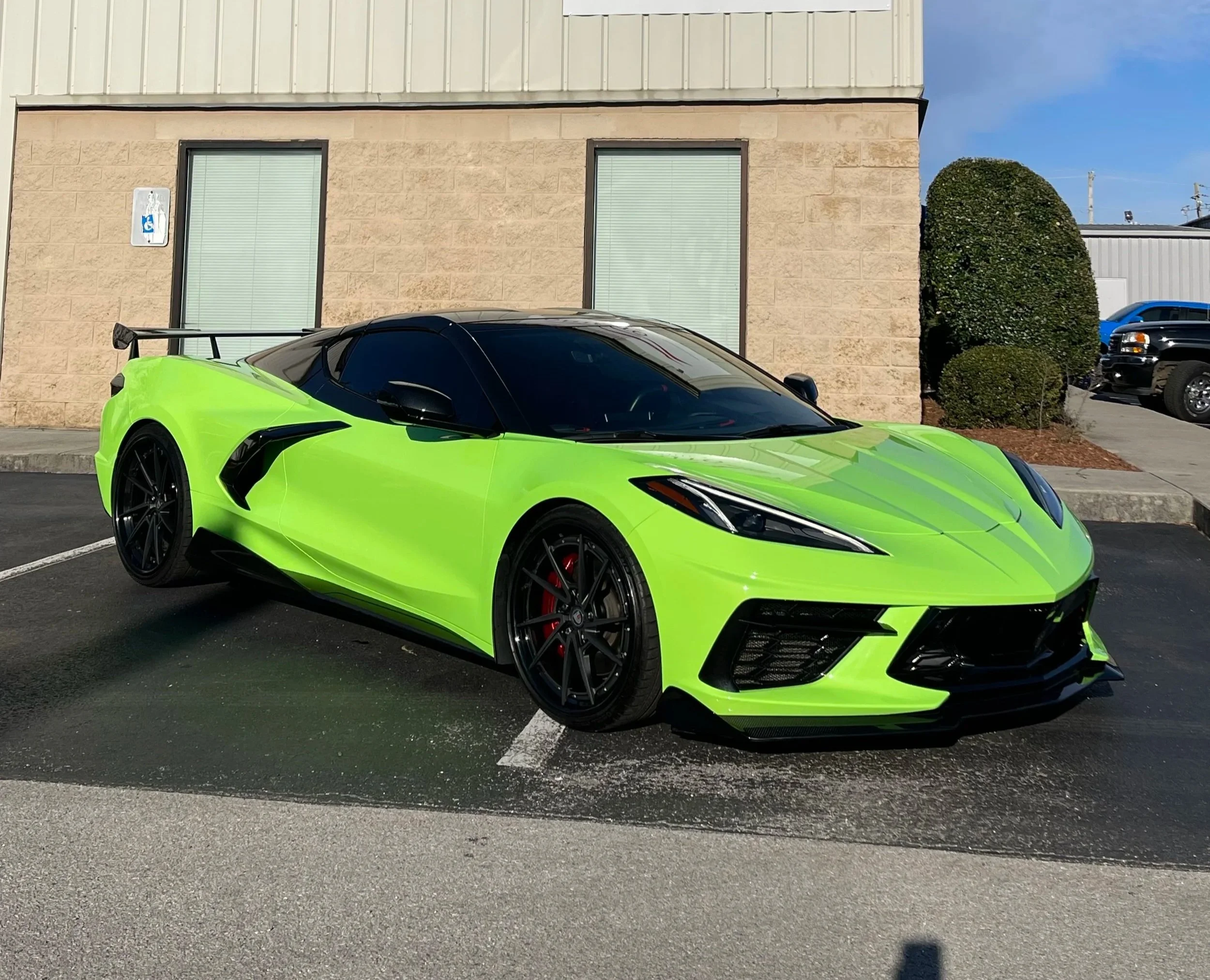 Bright green sports car with a black roof and wheels parked in a lot, with a building and bushes in the background.