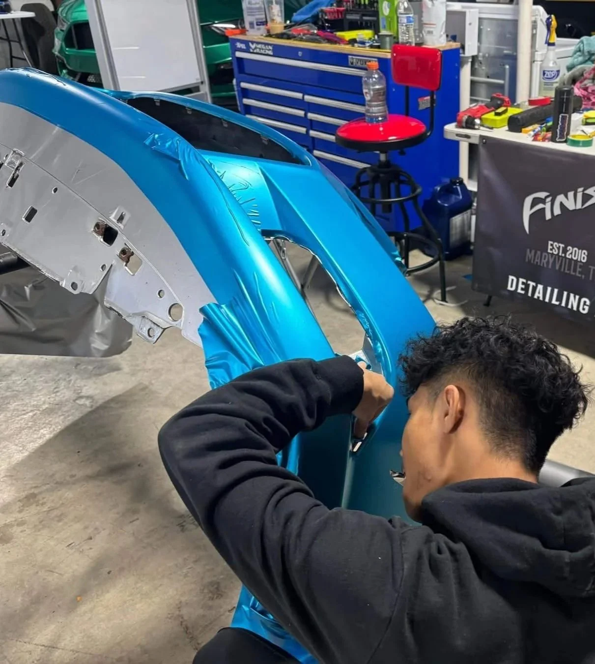 A person is working on a blue car part in a workshop, surrounded by tools and equipment, with a blue tool chest and a red stool in the background.