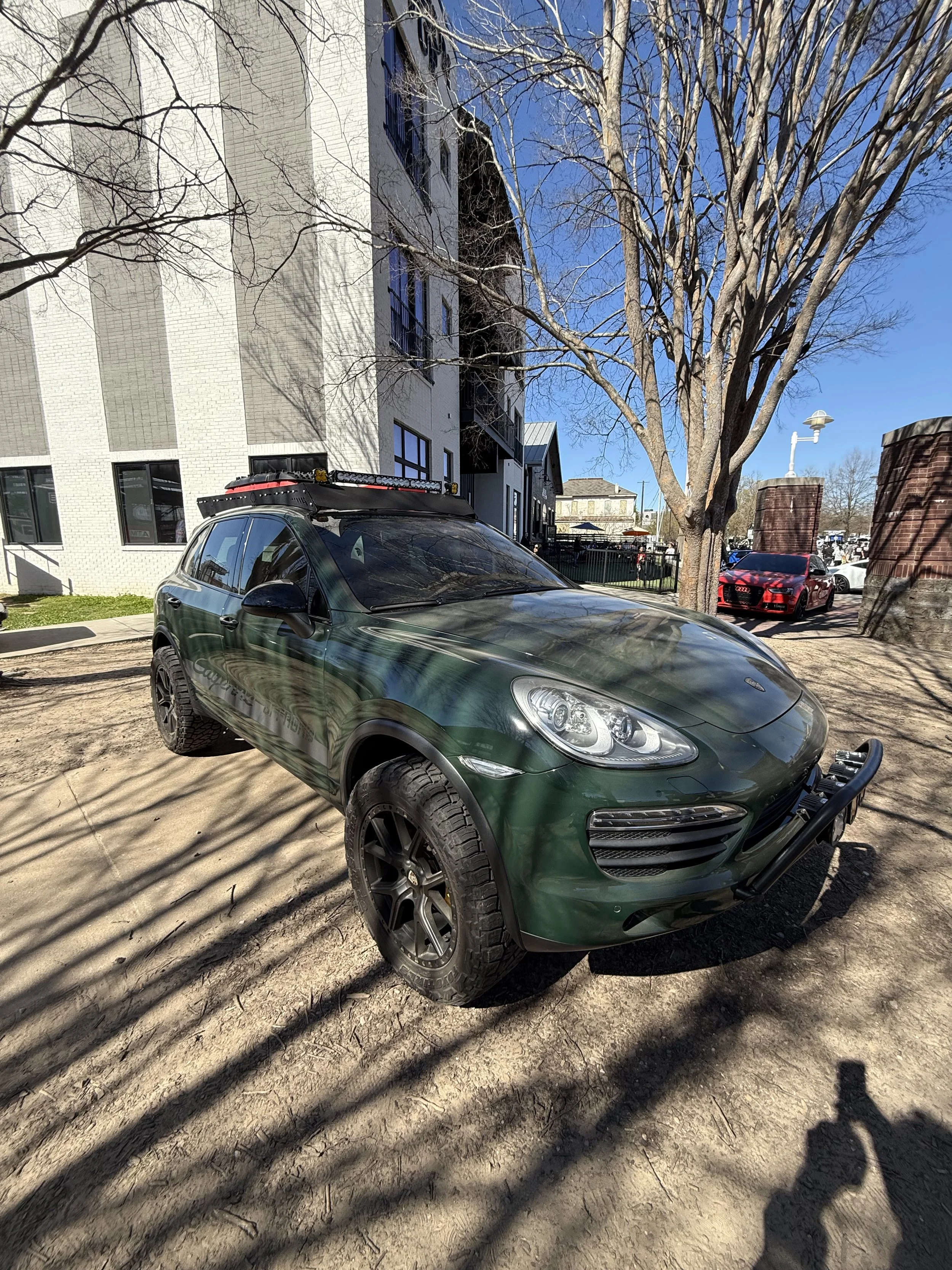 A green Porsche SUV parked outdoors on a dirt surface next to leafless trees and a white building, with other cars and buildings in the background.