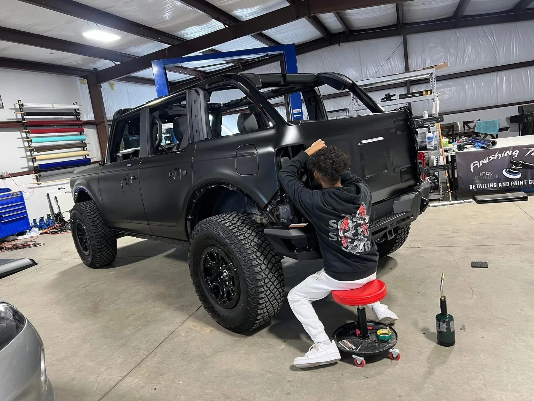 A person working on customizing a matte black off-road SUV inside a garage or workshop, sitting on a red rolling stool, with tools and equipment around.