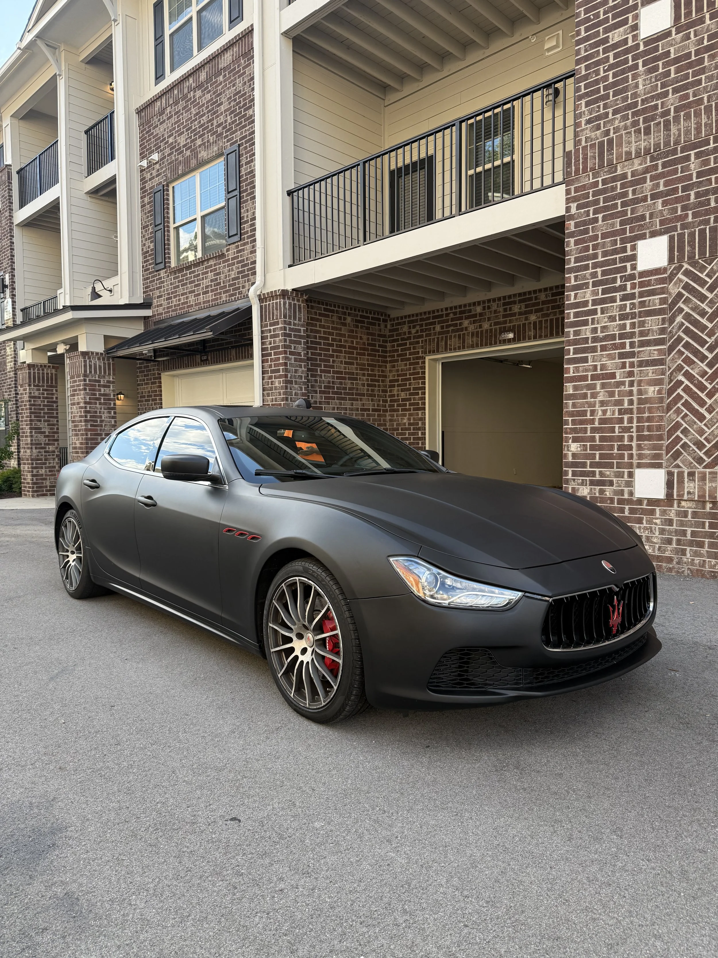 A matte black Maserati sports car is parked on a residential street in front of a multi-story brick apartment building. The car has distinctive Maserati logo on the front grille, red brake calipers, and stylish multi-spoke wheels.