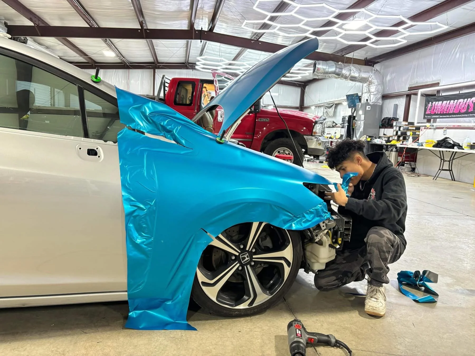 A person working on installing blue wrap on a white car in an indoor workshop, with a red truck and workshop tools in the background.
