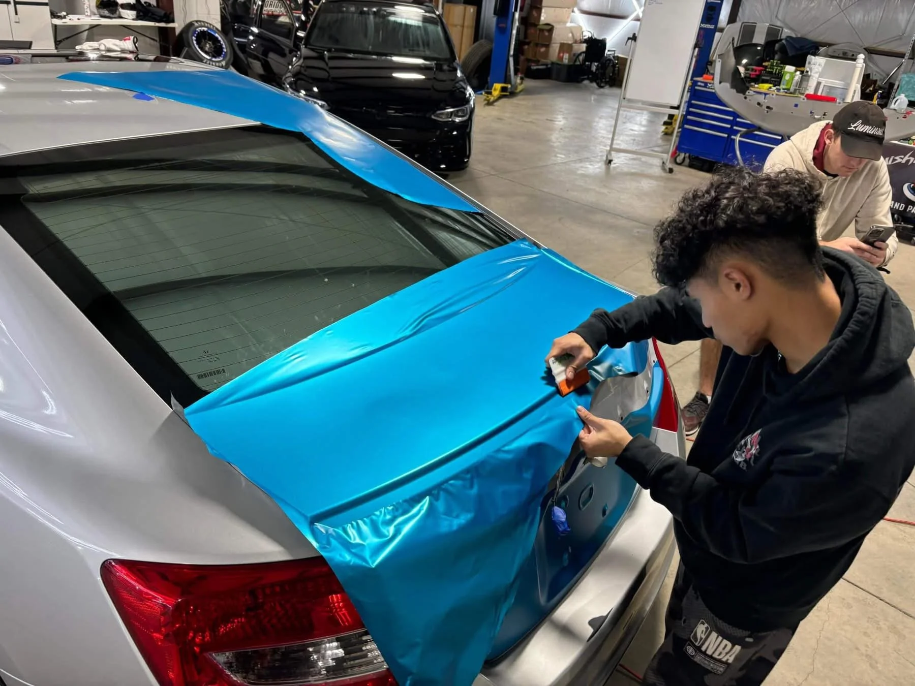A young man in a black hoodie applying blue vinyl wrap to the trunk of a silver car in a workshop, with another person in the background using a phone.