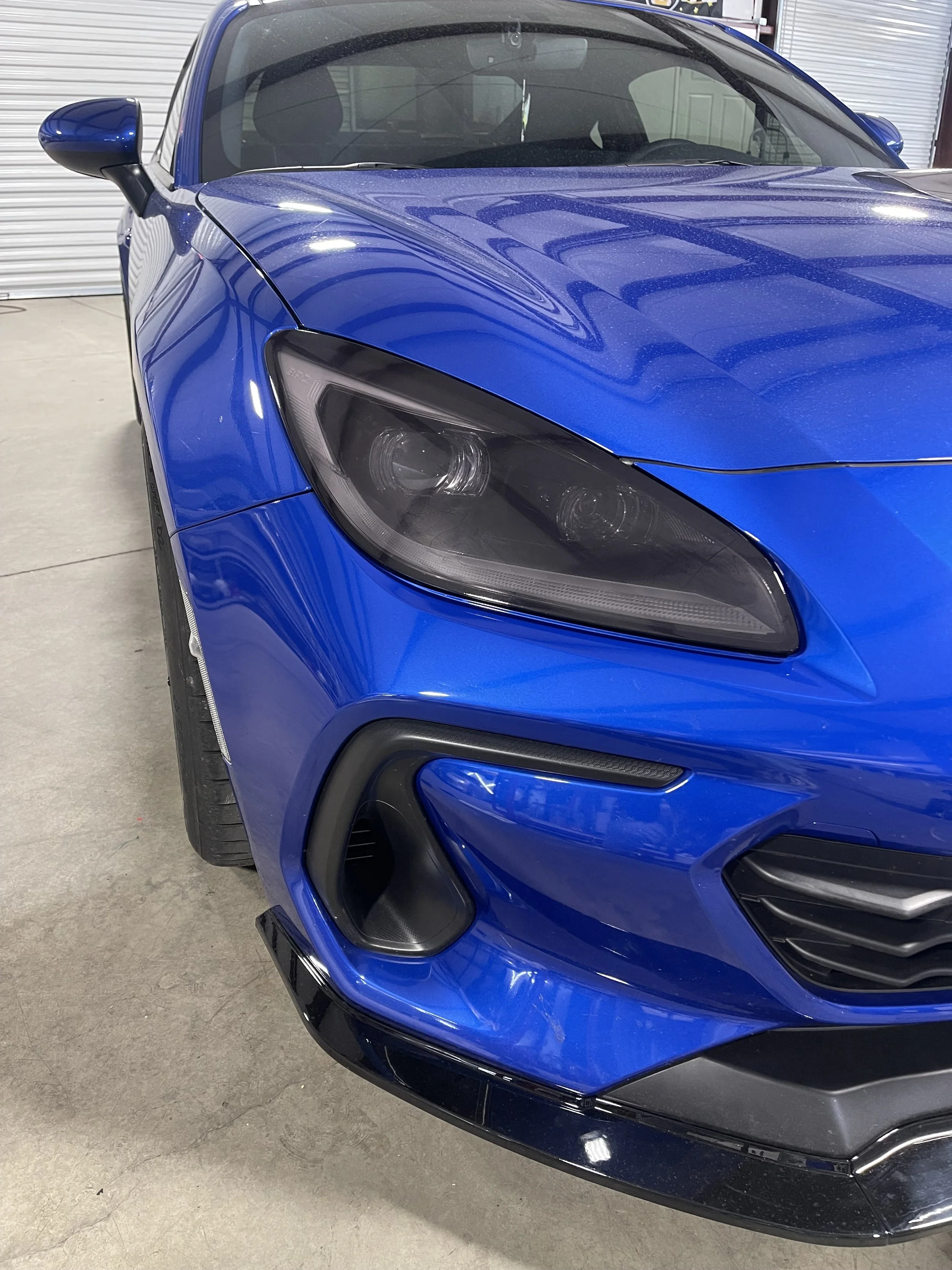 Close-up of the front left side of a blue sports car, showing the headlight, front bumper, and a wheel in a garage with a concrete floor and white rolling door in the background.