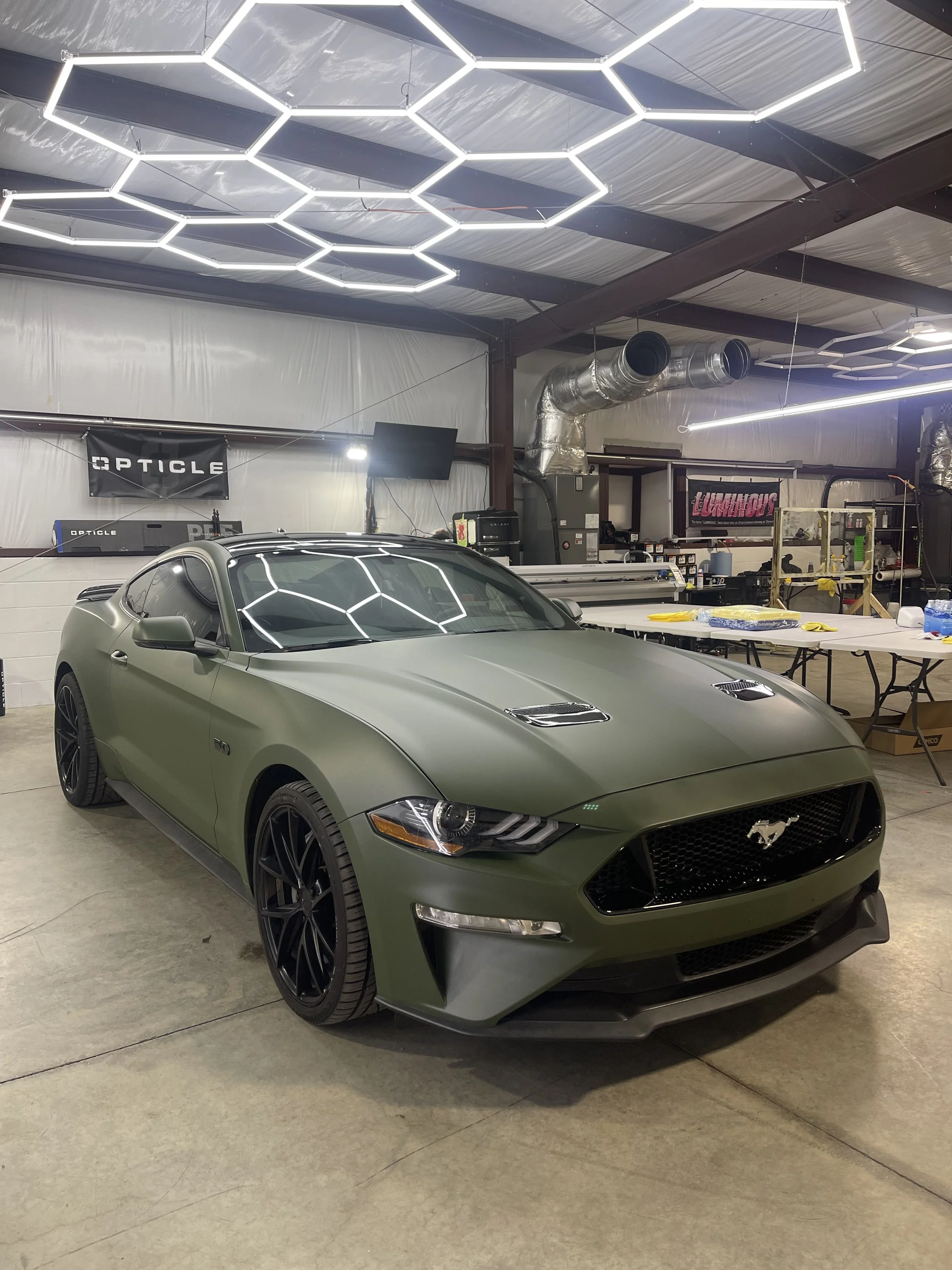 A matte green Ford Mustang with black wheels parked inside a garage or workshop with hexagonal LED ceiling lights.