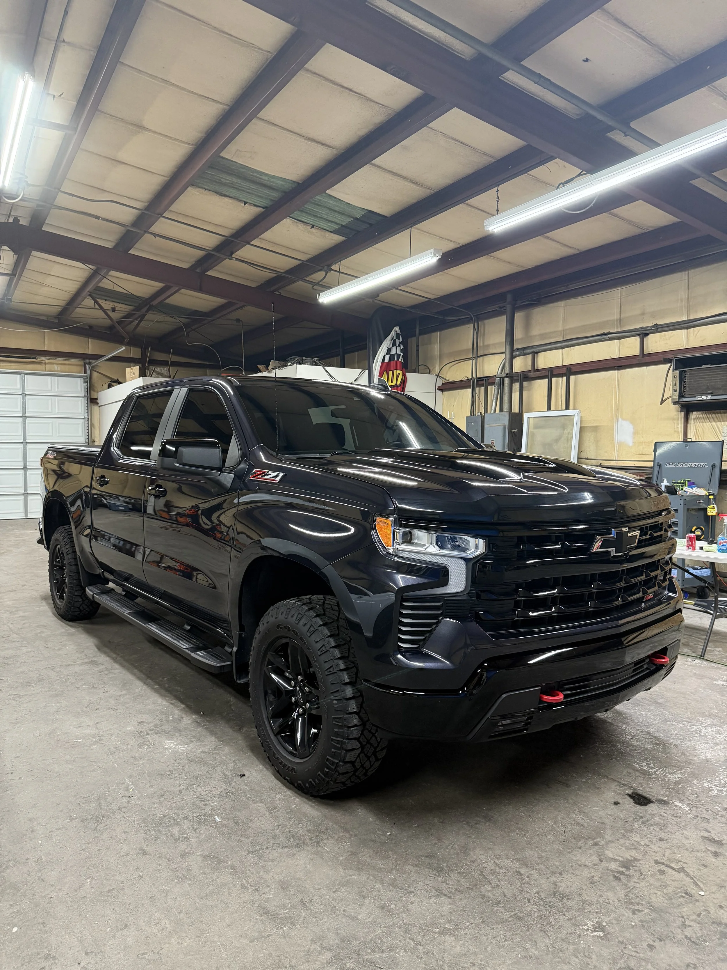 A black Chevrolet Silverado Z71 pickup truck is parked inside a garage with a concrete floor and a high ceiling, surrounded by tools and equipment.