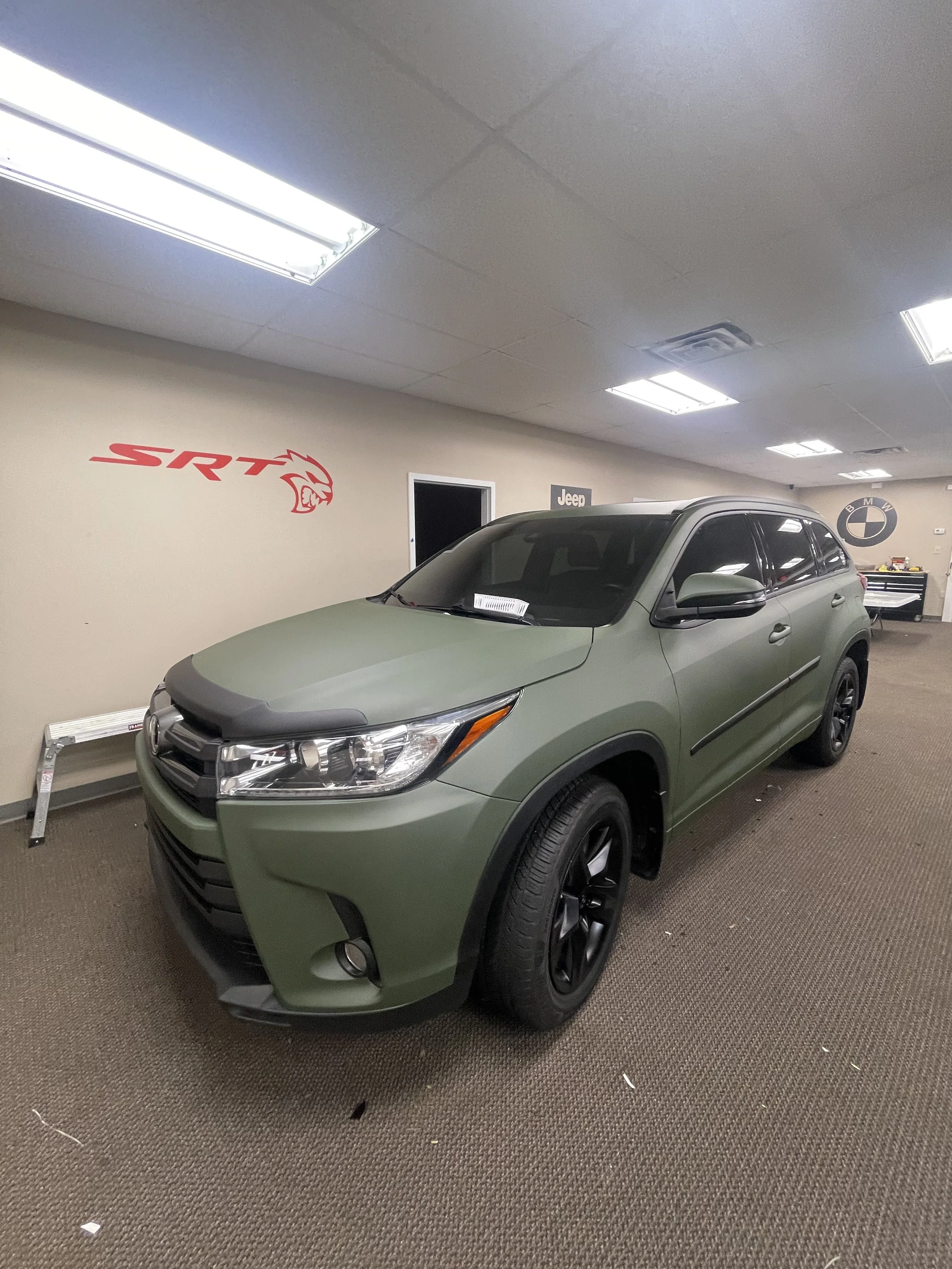 A green Honda SUV is parked indoors in front of a beige wall with automotive brand logos, including 'SRT', 'Jeep', and 'BMW'.