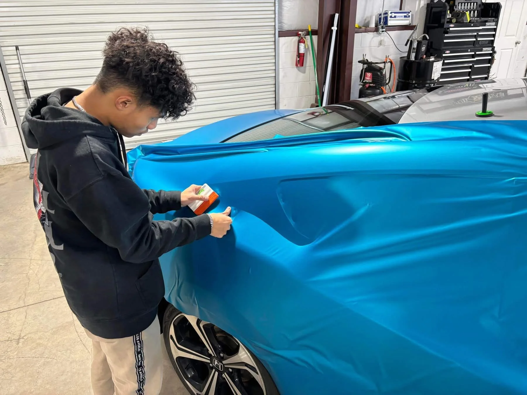 A person applying blue wrap film to a sports car in a garage.