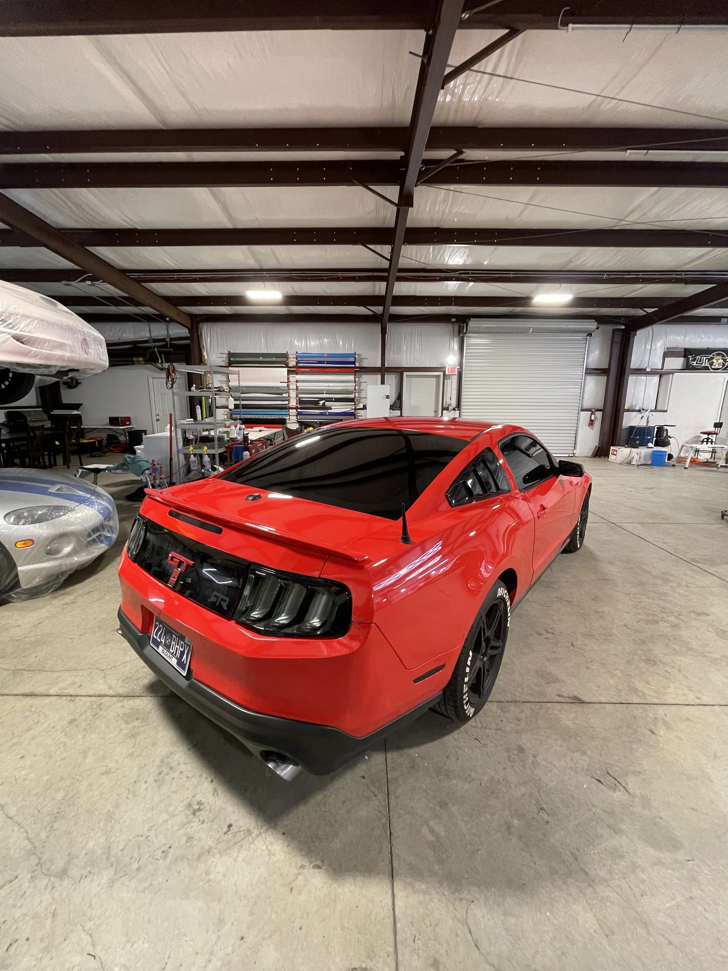 A red Ford Mustang GT parked inside a spacious garage with a high ceiling and exposed beams. The car has tinted windows, black wheels, and a black rear spoiler. In the background, there are shelves with car wraps and various tools and equipment.