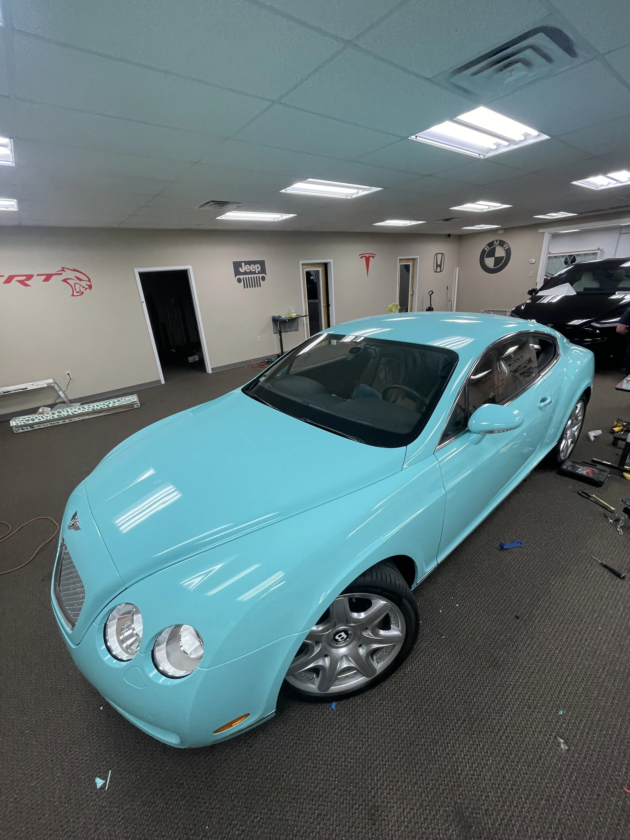 A light blue luxury sports car parked inside a showroom or garage with car brand logos on the wall in the background.