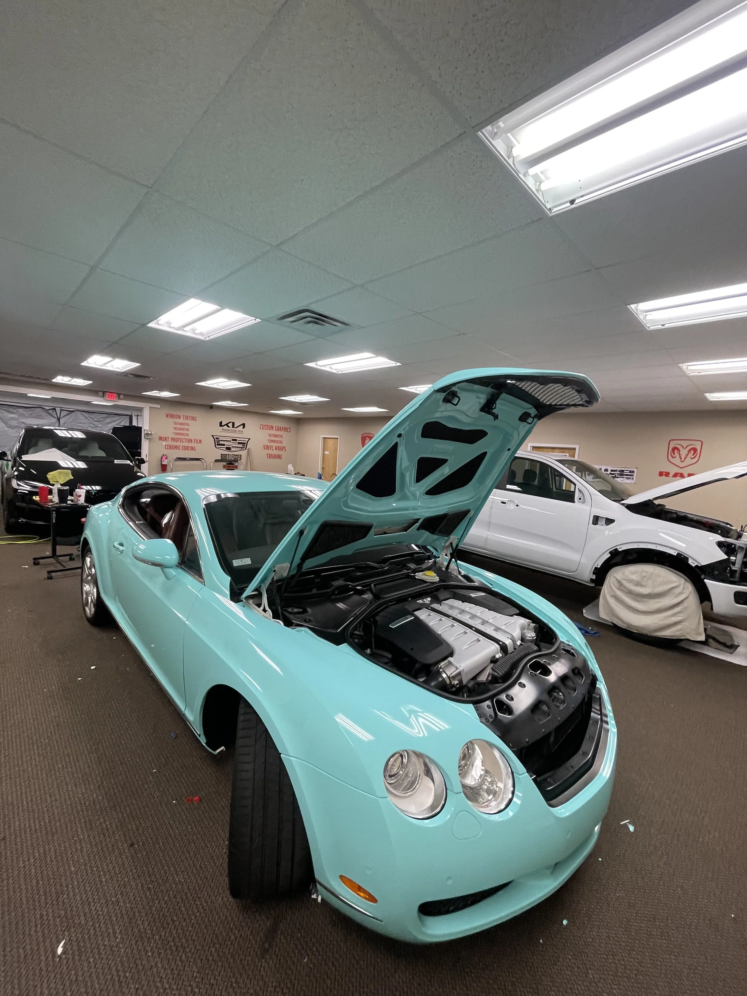 Light blue sports car with open hood inside an auto shop, surrounded by other vehicles and car care equipment.
