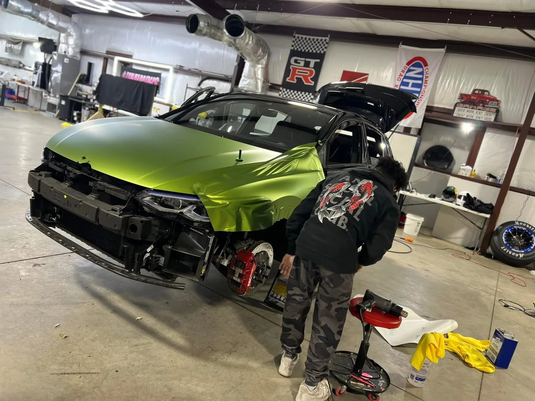A person working on a partially assembled car in a garage, with the car's front bumper and parts removed, revealing the suspension and brake system, and a green wrap covering part of the hood.