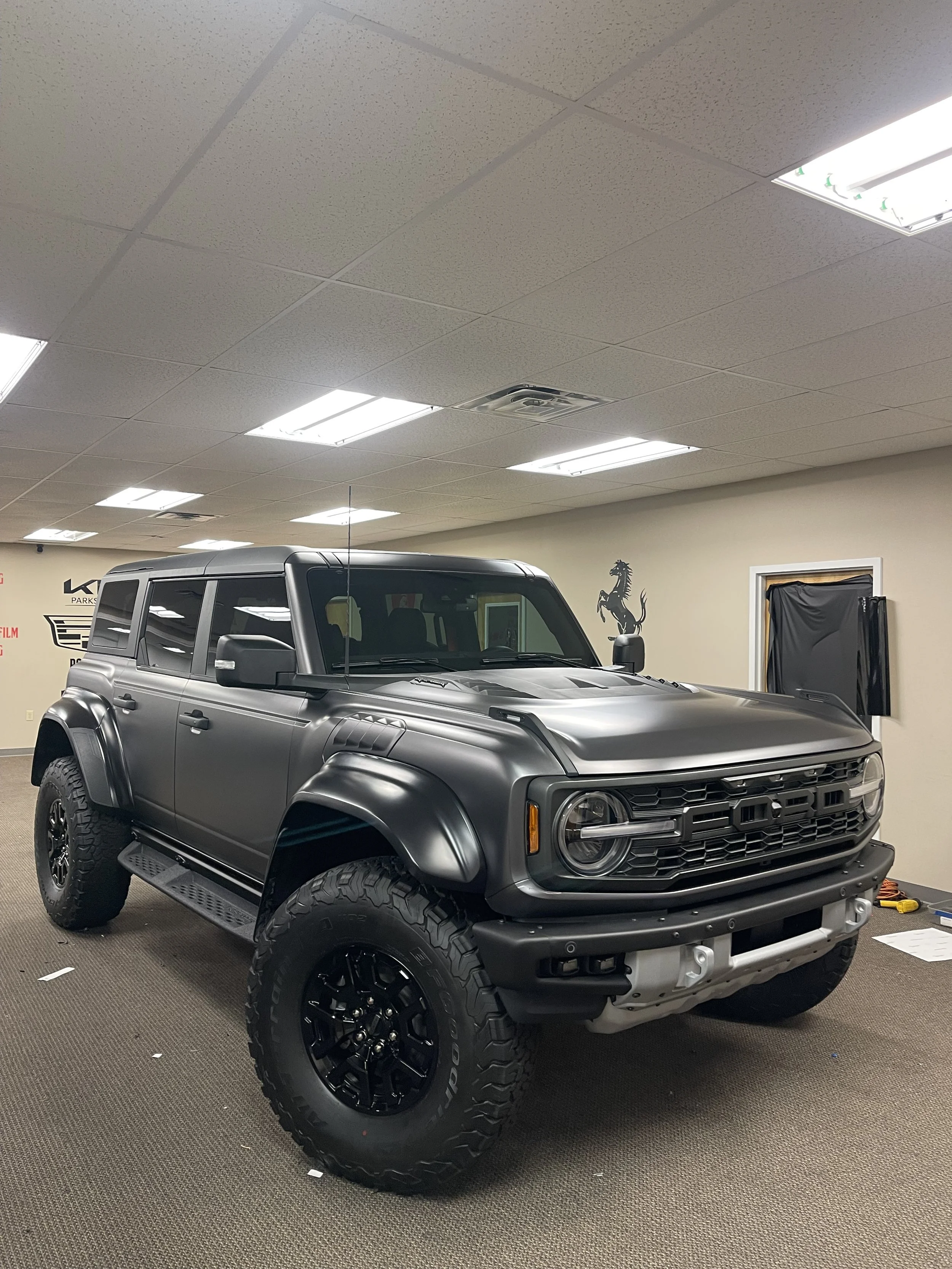 A black off-road vehicle in a room with white ceiling tiles and fluorescent lighting, featuring large tires, a matte black body, and a rugged design.