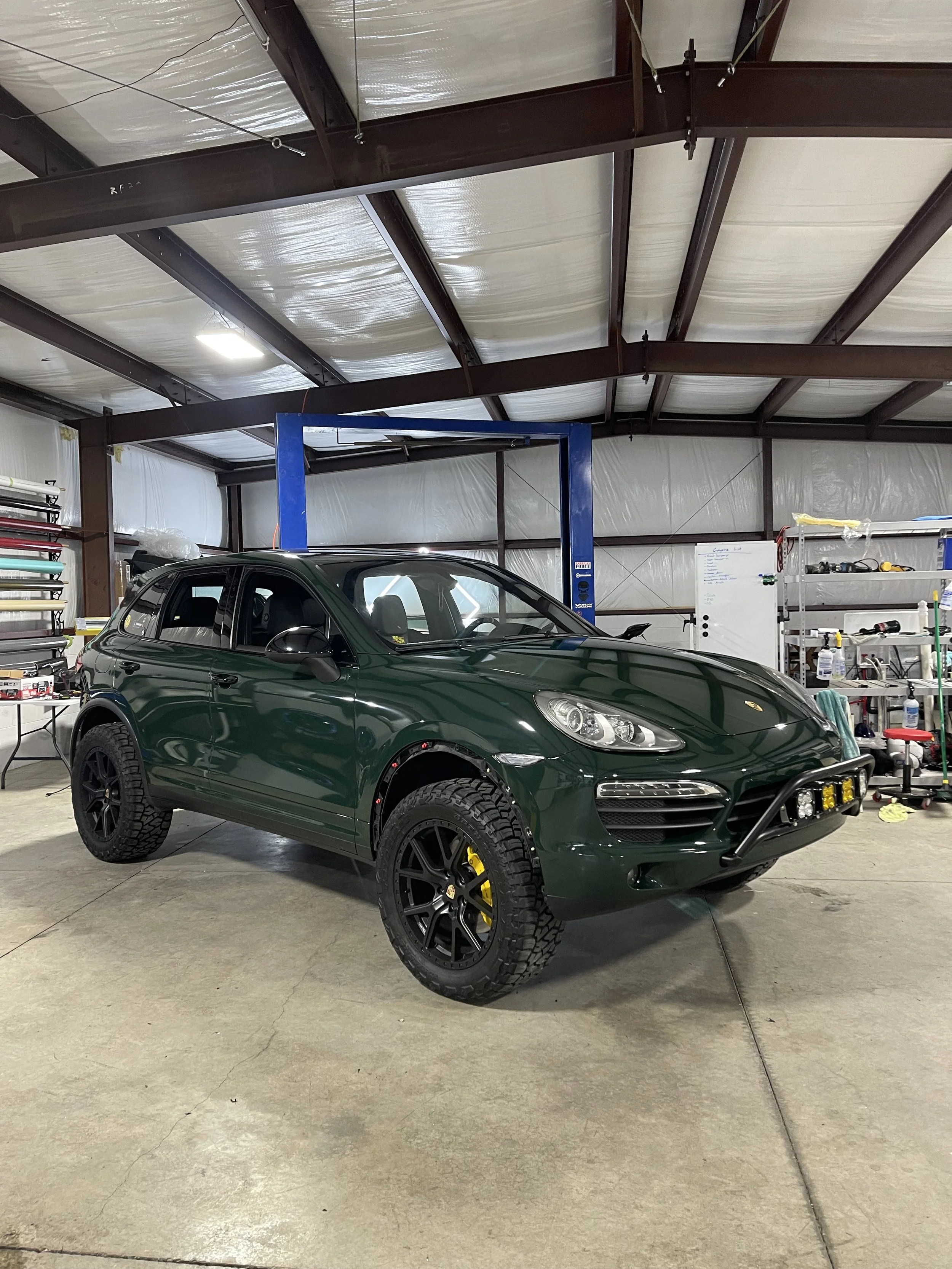 A green Porsche SUV inside a garage or workshop, with off-road tires, black rims, yellow brake calipers, and a front bull bar with lights.