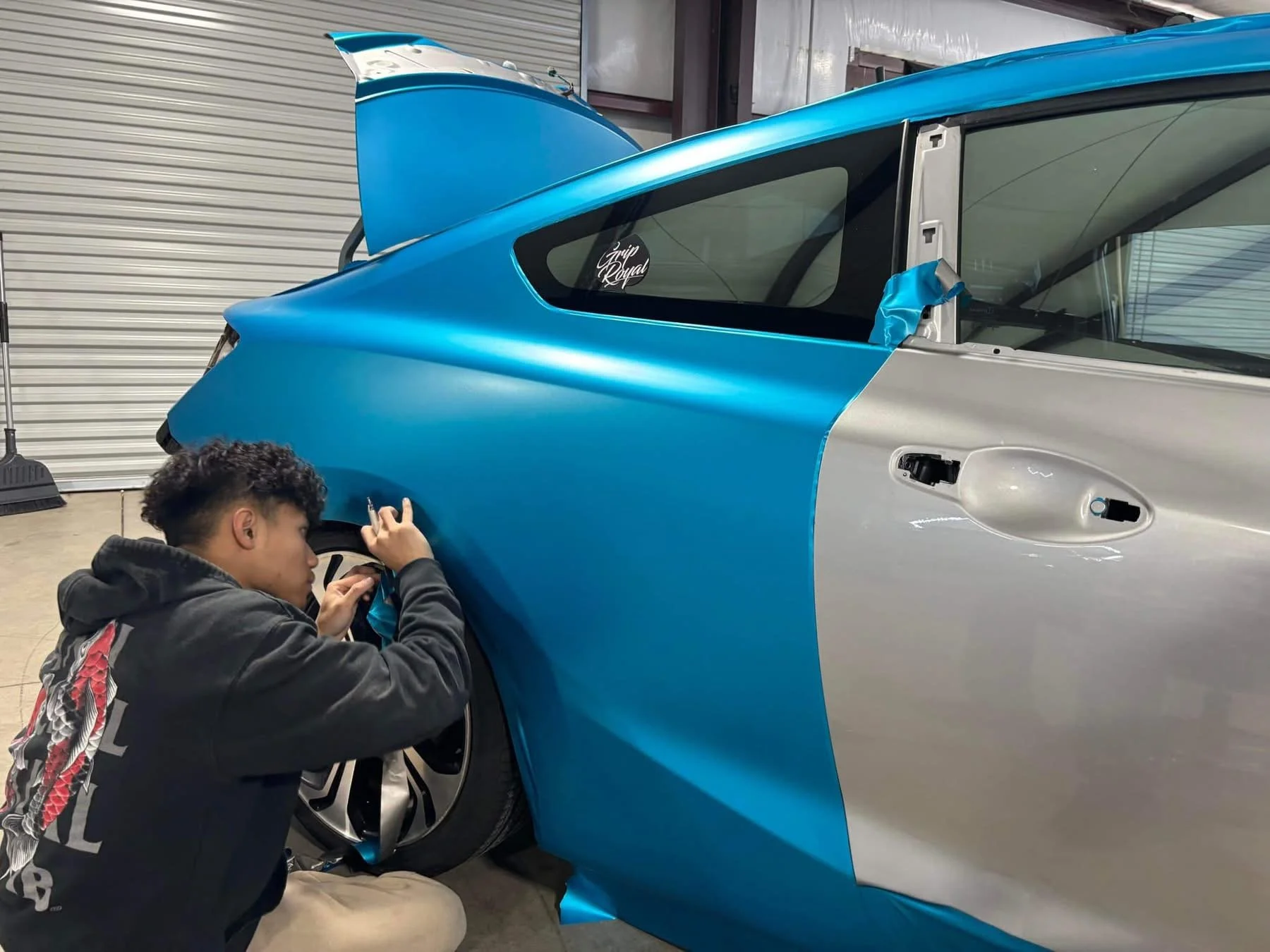 Person working on a blue and silver sports car, applying paint or decals in a garage or workshop.