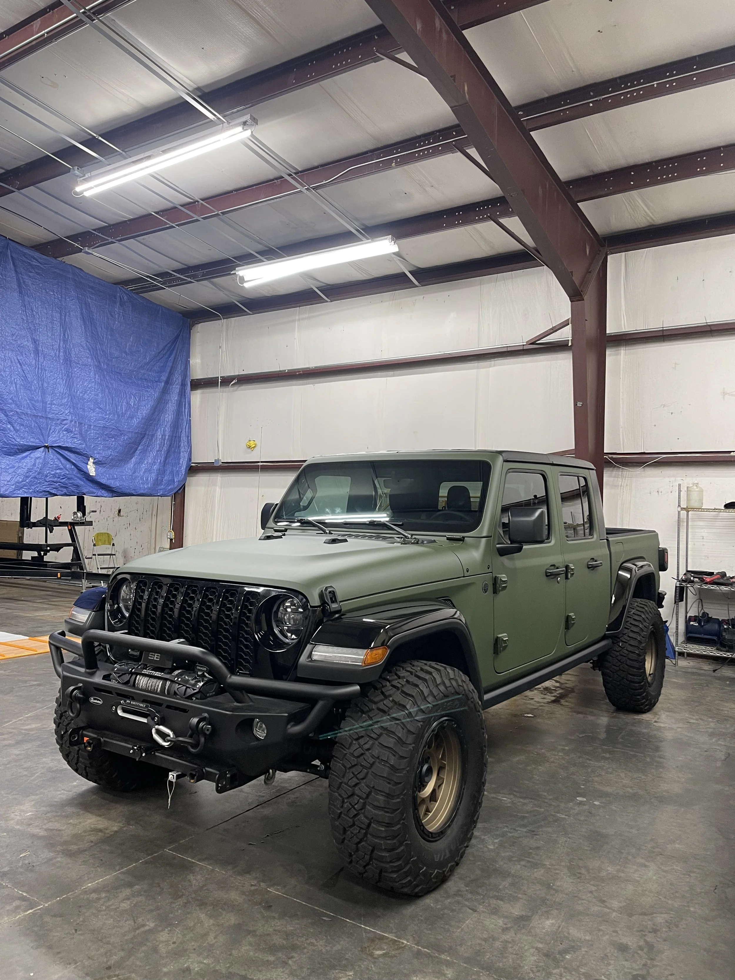 A green, four-door, four-wheel drive Jeep with off-road tires and a heavy-duty front bumper, parked inside a large industrial garage with metal beams, fluorescent lights, and a blue tarp covering part of the wall.