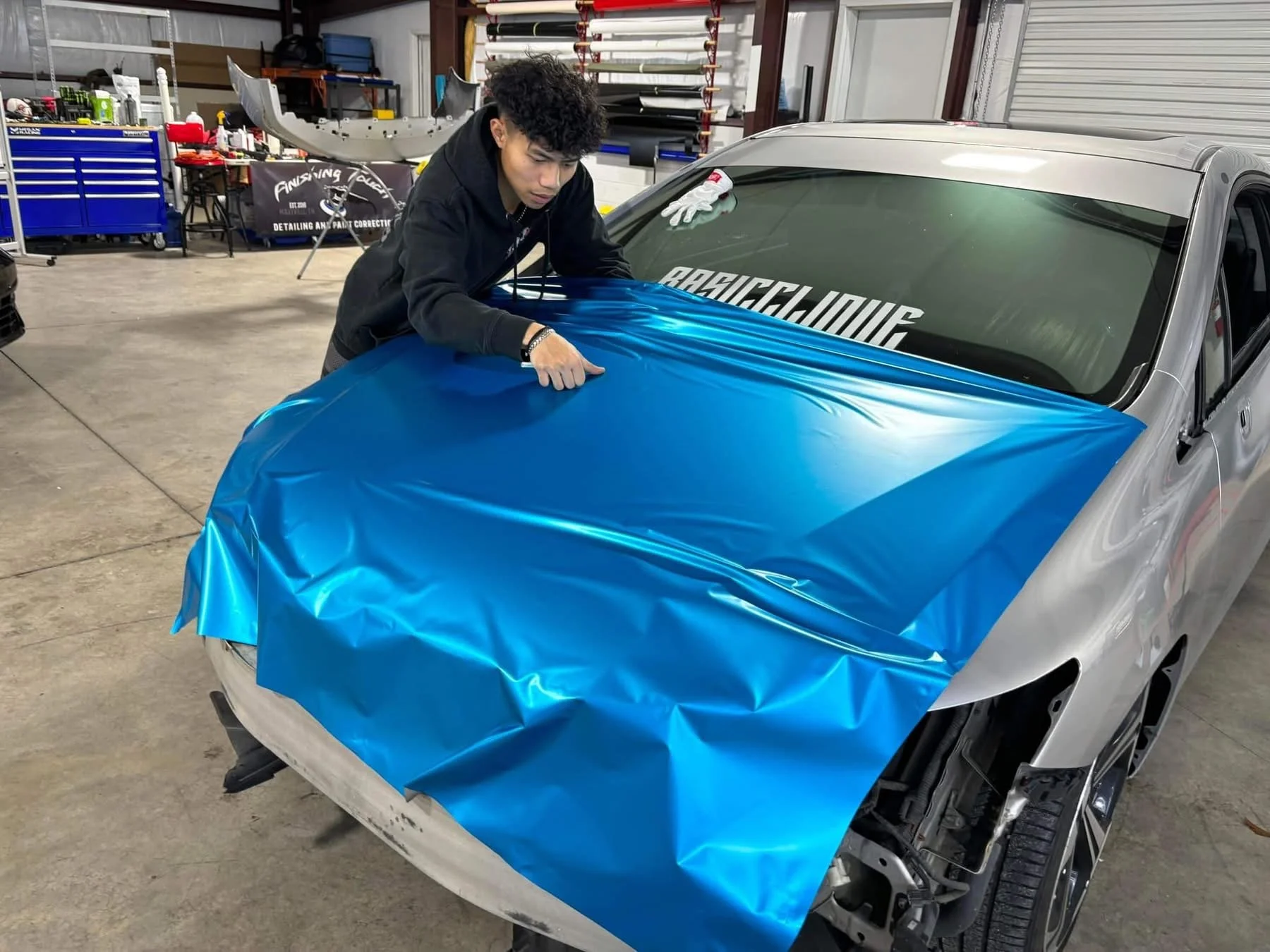 A person applying blue vinyl wrap to the hood of a silver car inside a workshop.