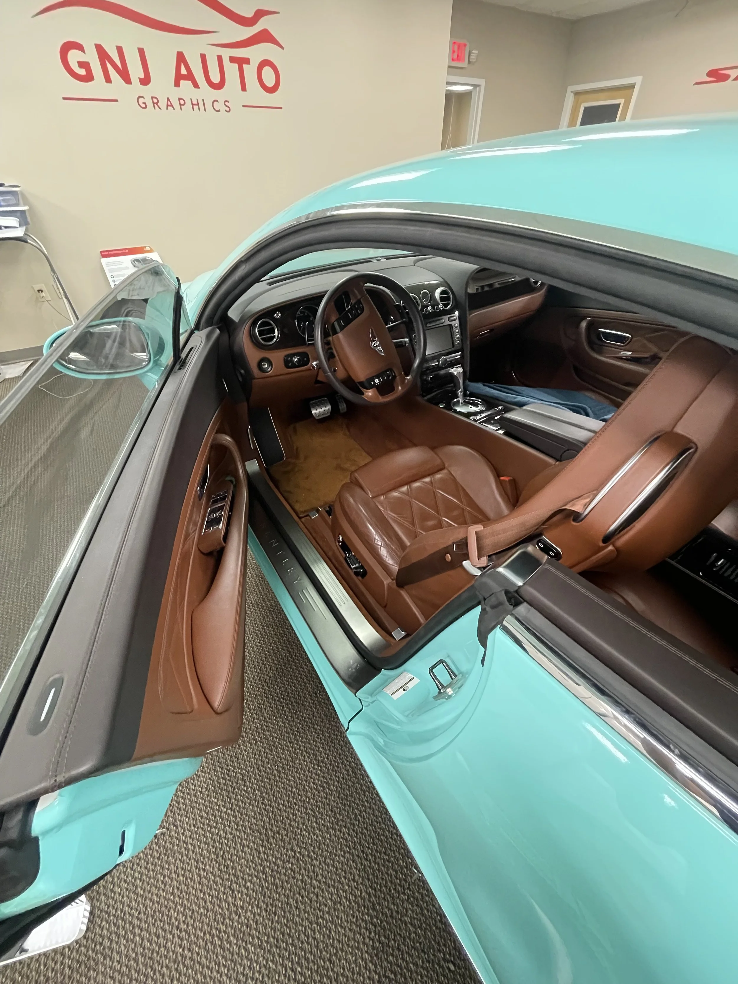 Interior of a vintage turquoise car with brown leather seats, dashboard, and steering wheel, displayed indoors at GNJ Auto Graphics.